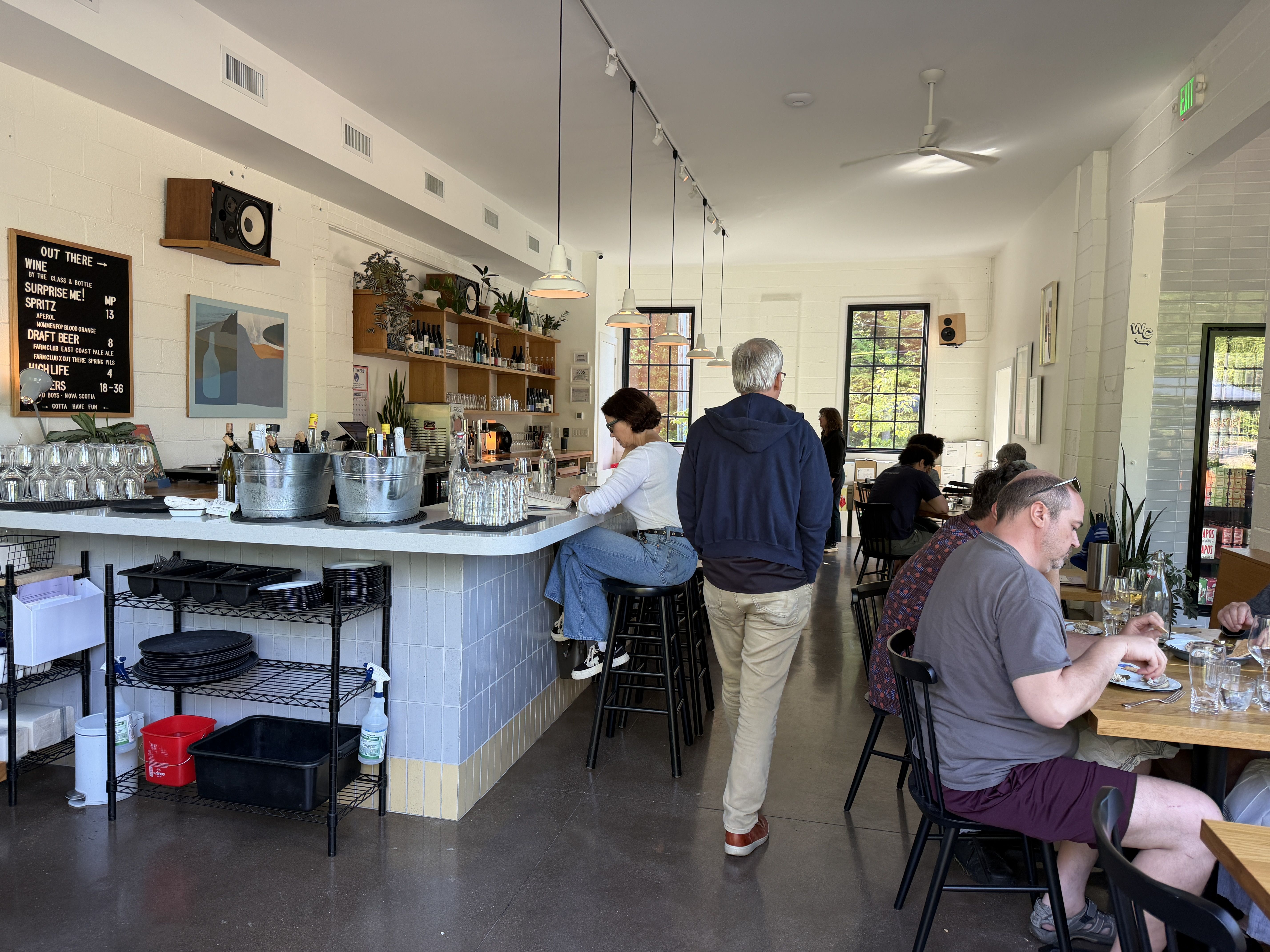 Bright cafe interior with a long white counter, hanging pendant lights, shelves of bottles and plants behind the bar, customers seated at tables, glassware on the counter, and a ceiling fan overhead.