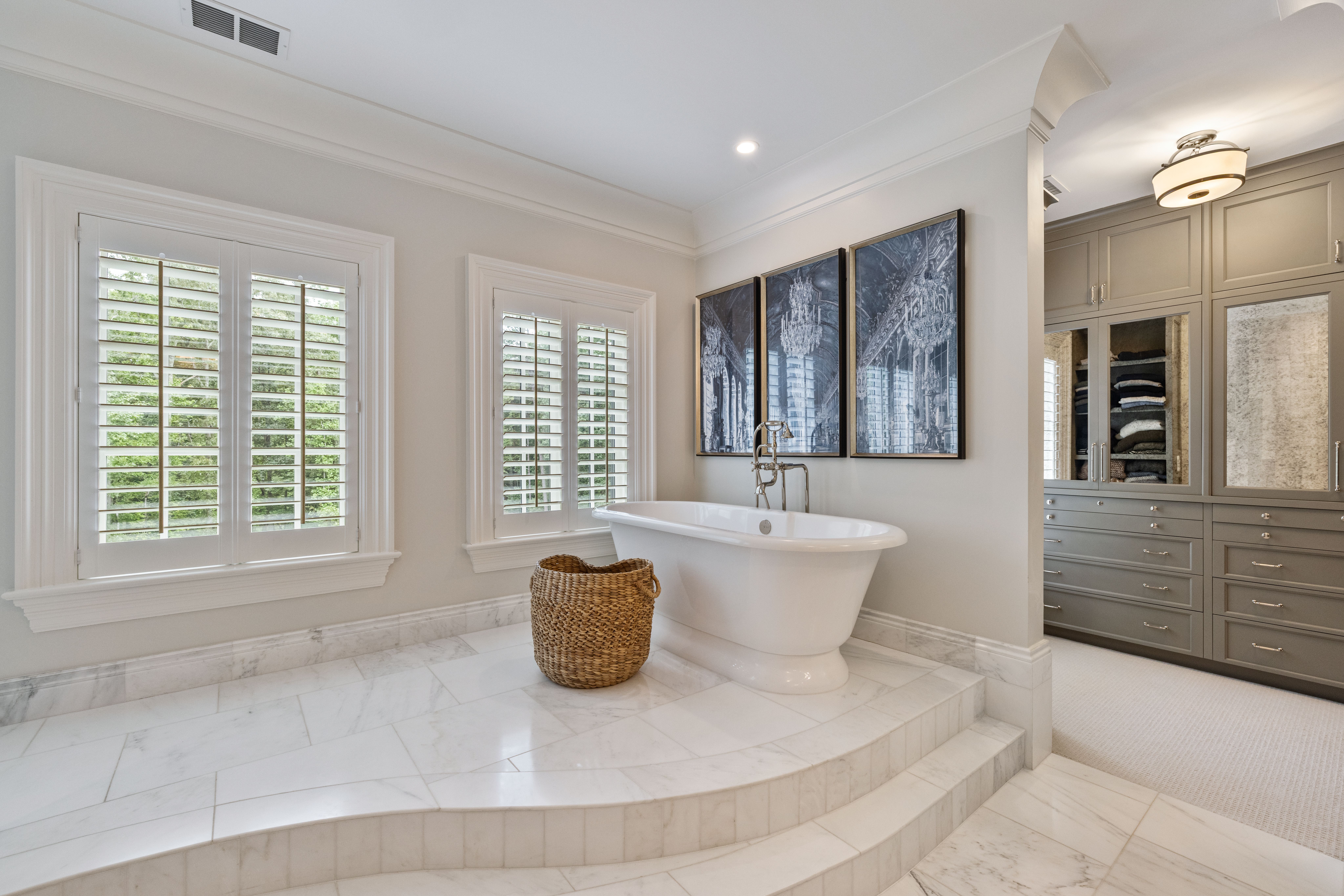 Elegant bathroom with white marble flooring, a white freestanding tub, a wicker basket, two shuttered windows, and three framed black-and-white chandelier prints above the tub. Adjacent gray cabinetry.