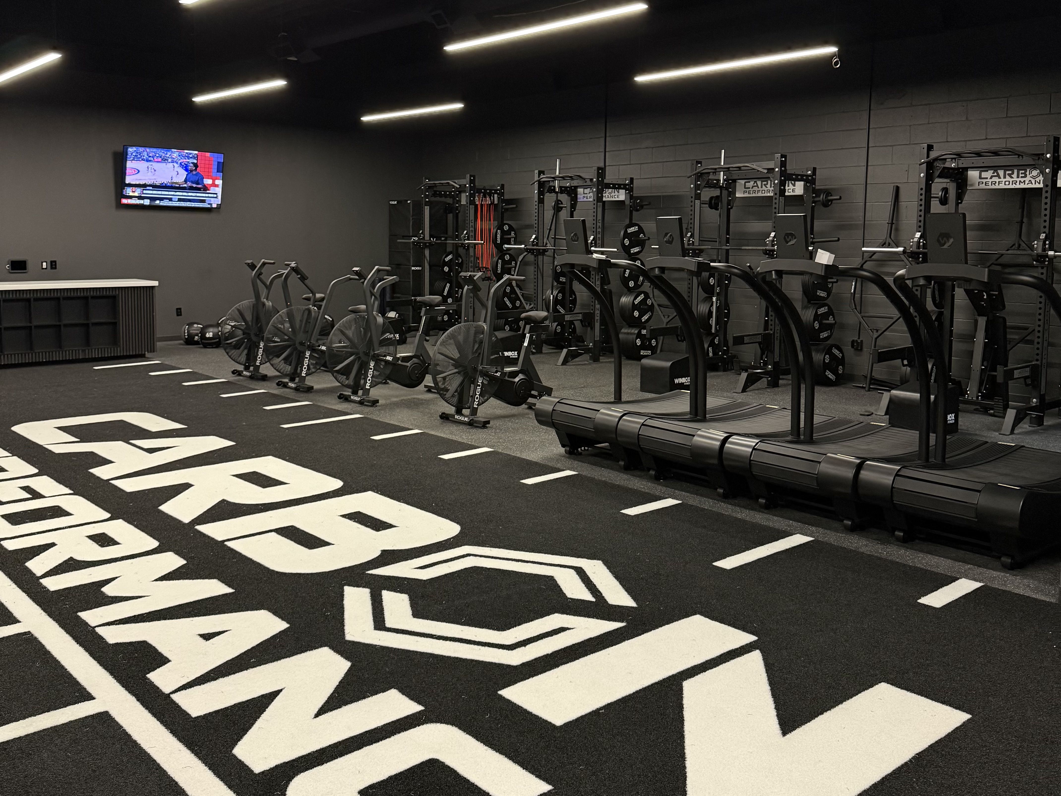 Industrial gym interior with black walls, bright linear lights, multiple squat racks, barbells and weight plates, cardio machines on left, a wall TV, and large white geometric floor graphics.