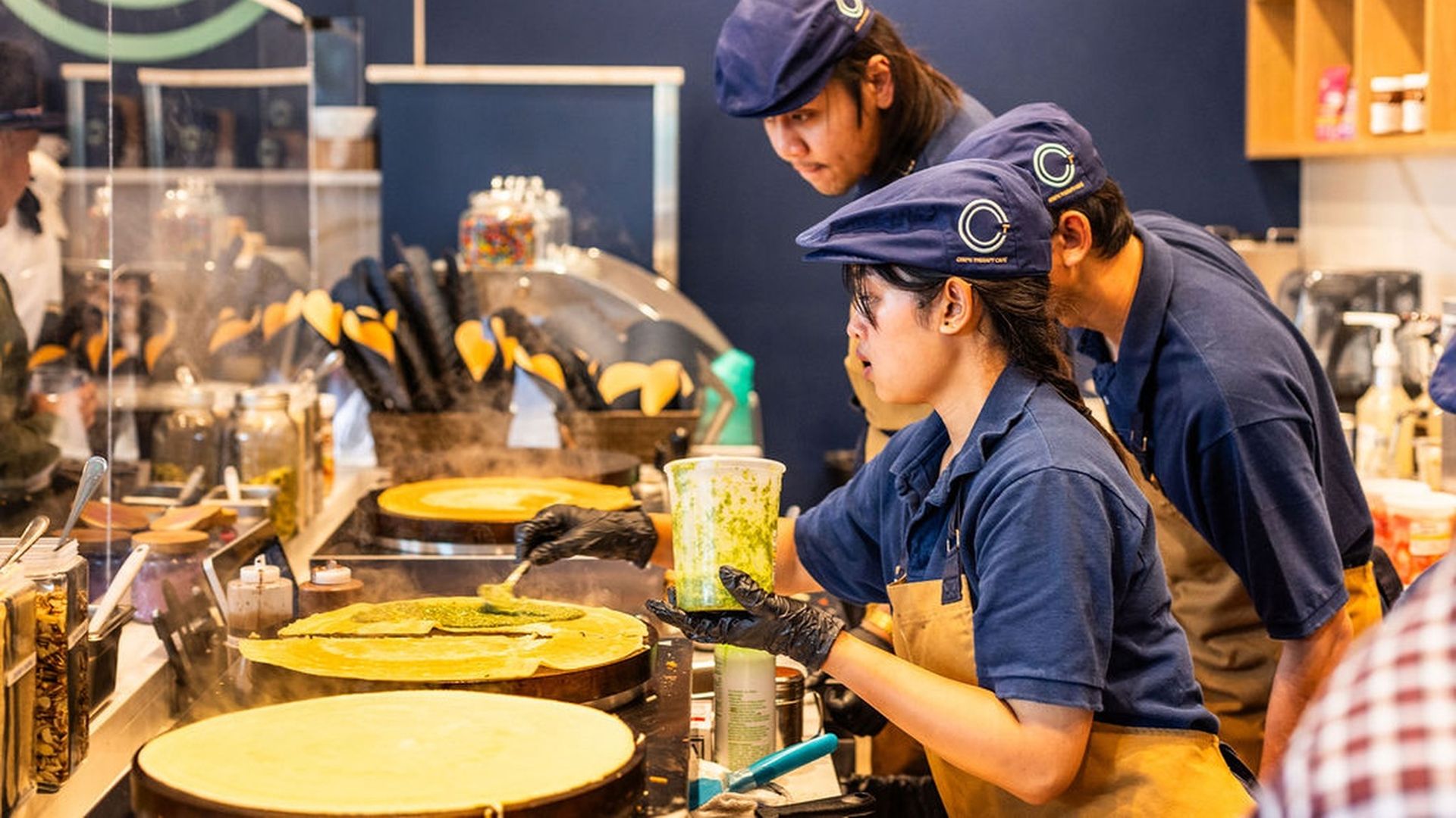 Three crepe shop workers in blue uniforms and tan aprons cook crepes at a busy counter; one pours green batter onto a crepe while two colleagues observe.