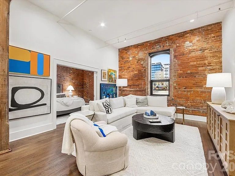 Bright living room with exposed red brick wall, white sectional sofa, beige armchair with blue pillow, black coffee table, modern art on white walls, and a window showing city buildings.