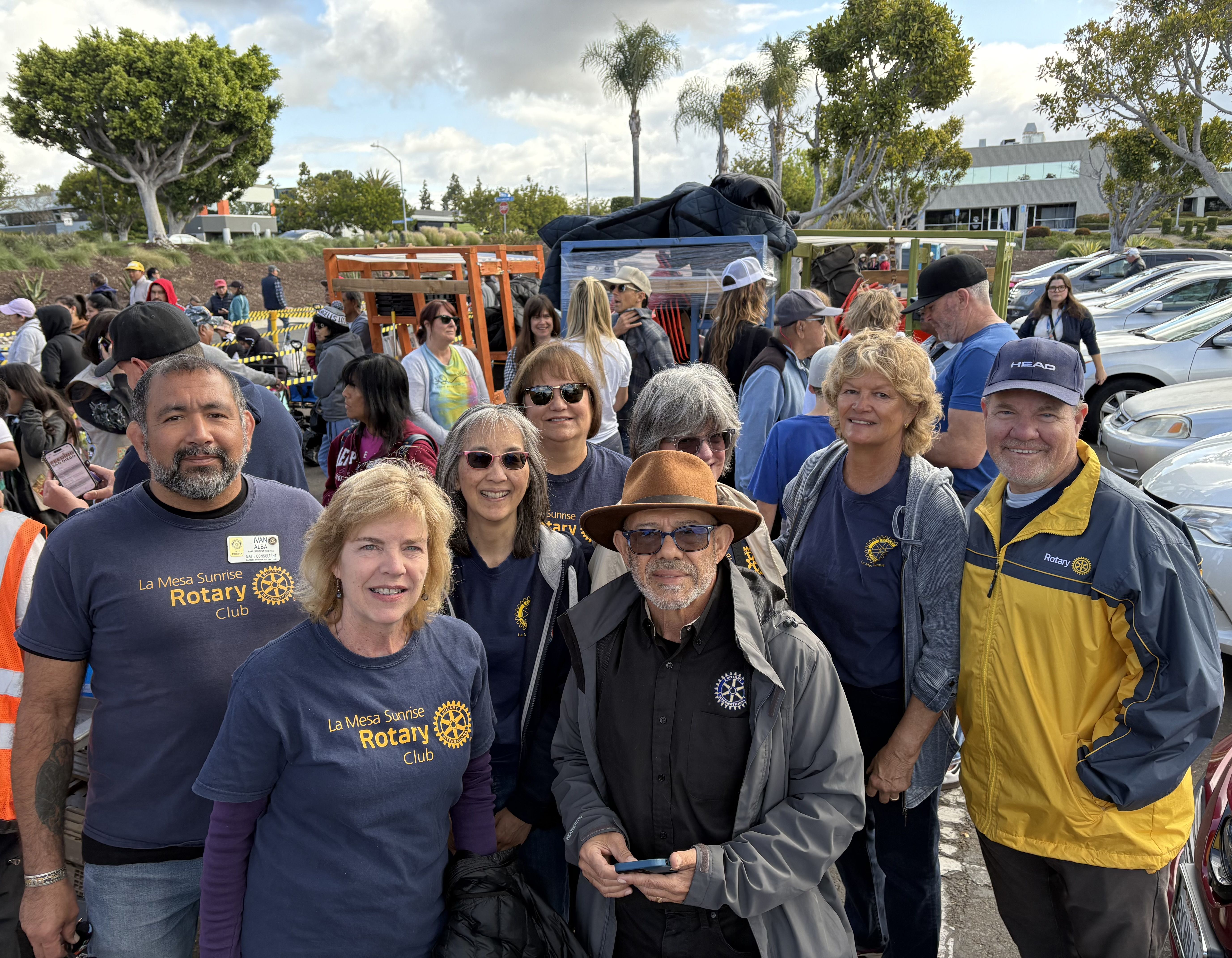 Crowd of smiling people at an outdoor Rotary event, many wearing navy shirts with Rotary Club logos; trees, a building, and parked cars visible in the background.