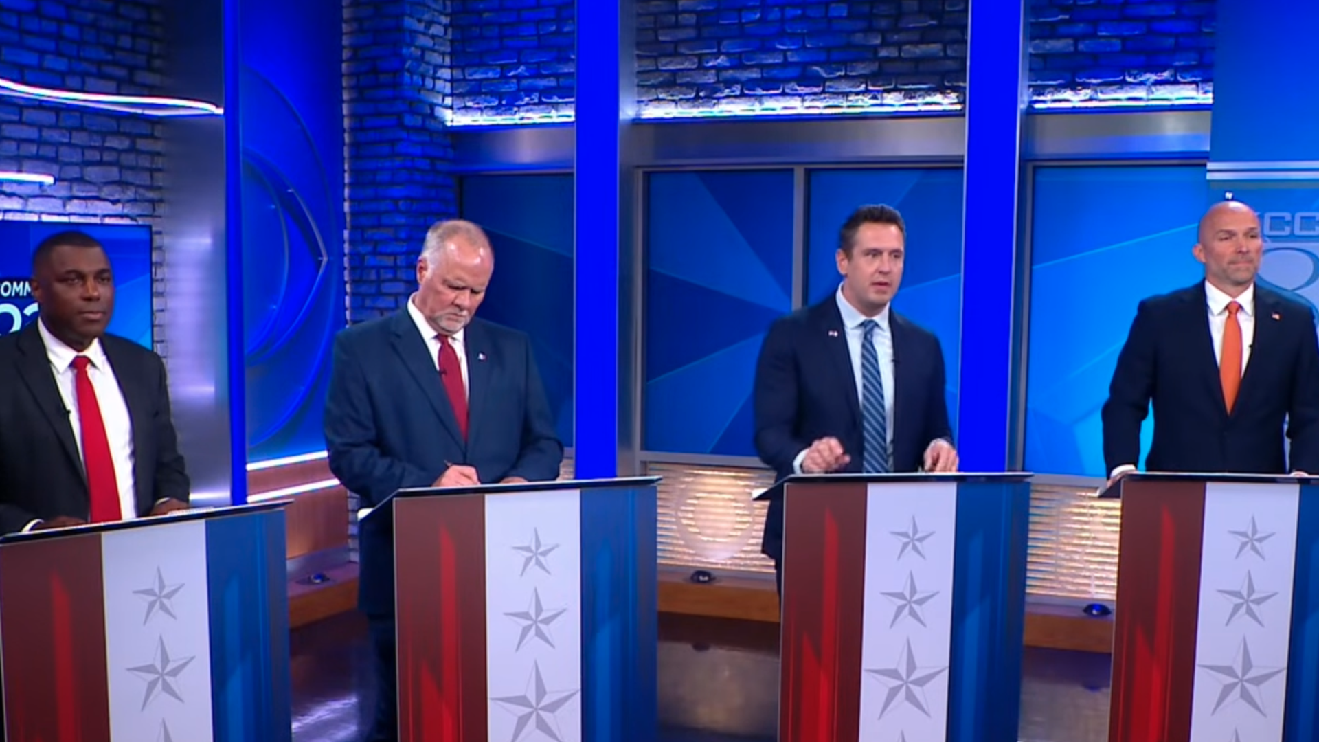 Four men in dark suits stand at podiums in a blue-lit TV debate studio; brick walls behind. Podiums feature star-pattern panels; ties are red, red, blue, and orange.