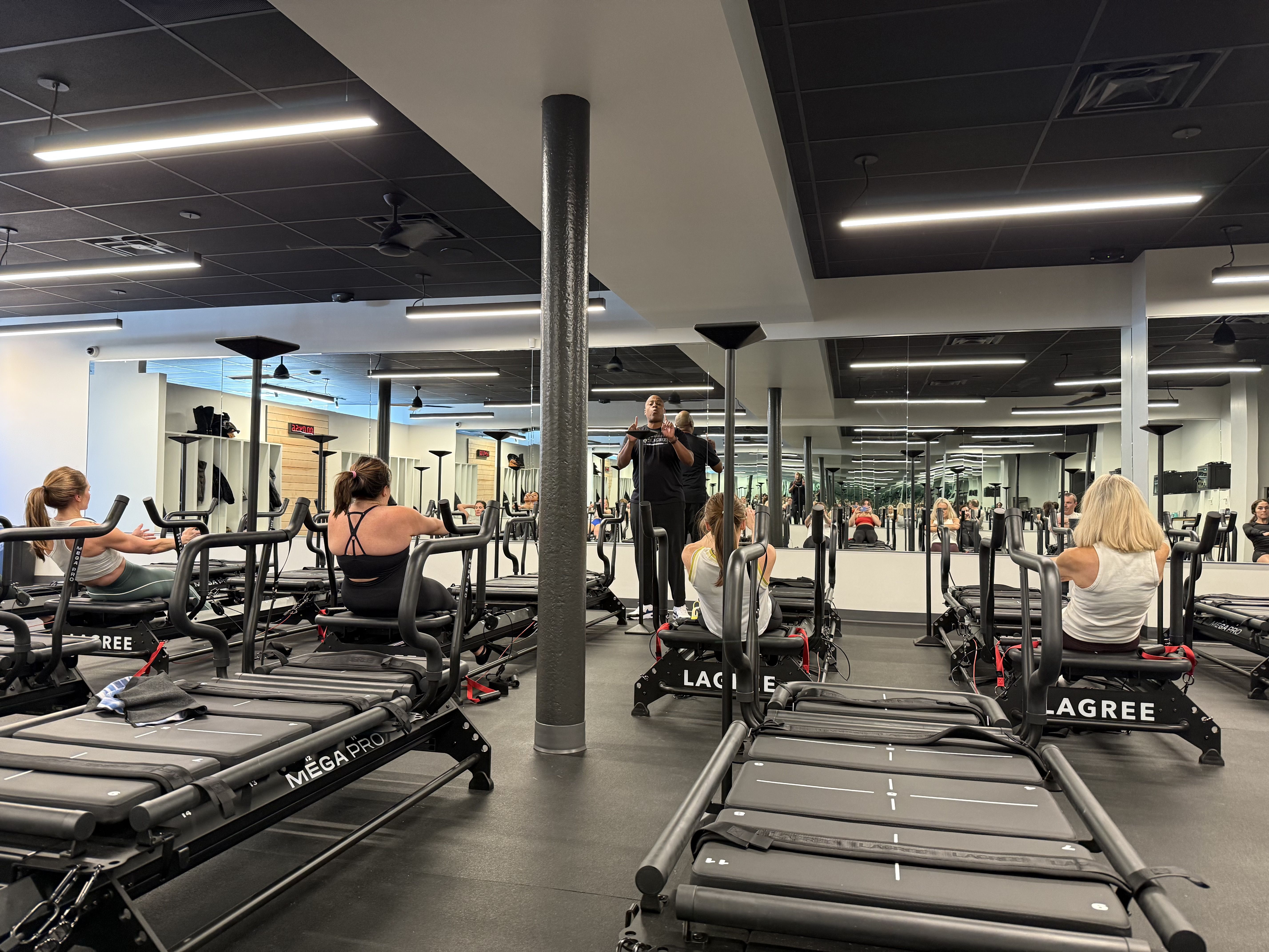 Four women on black Mega Pro Lagree fitness machines in a bright gym with black ceiling and large mirrors, following instruction from a standing male trainer.