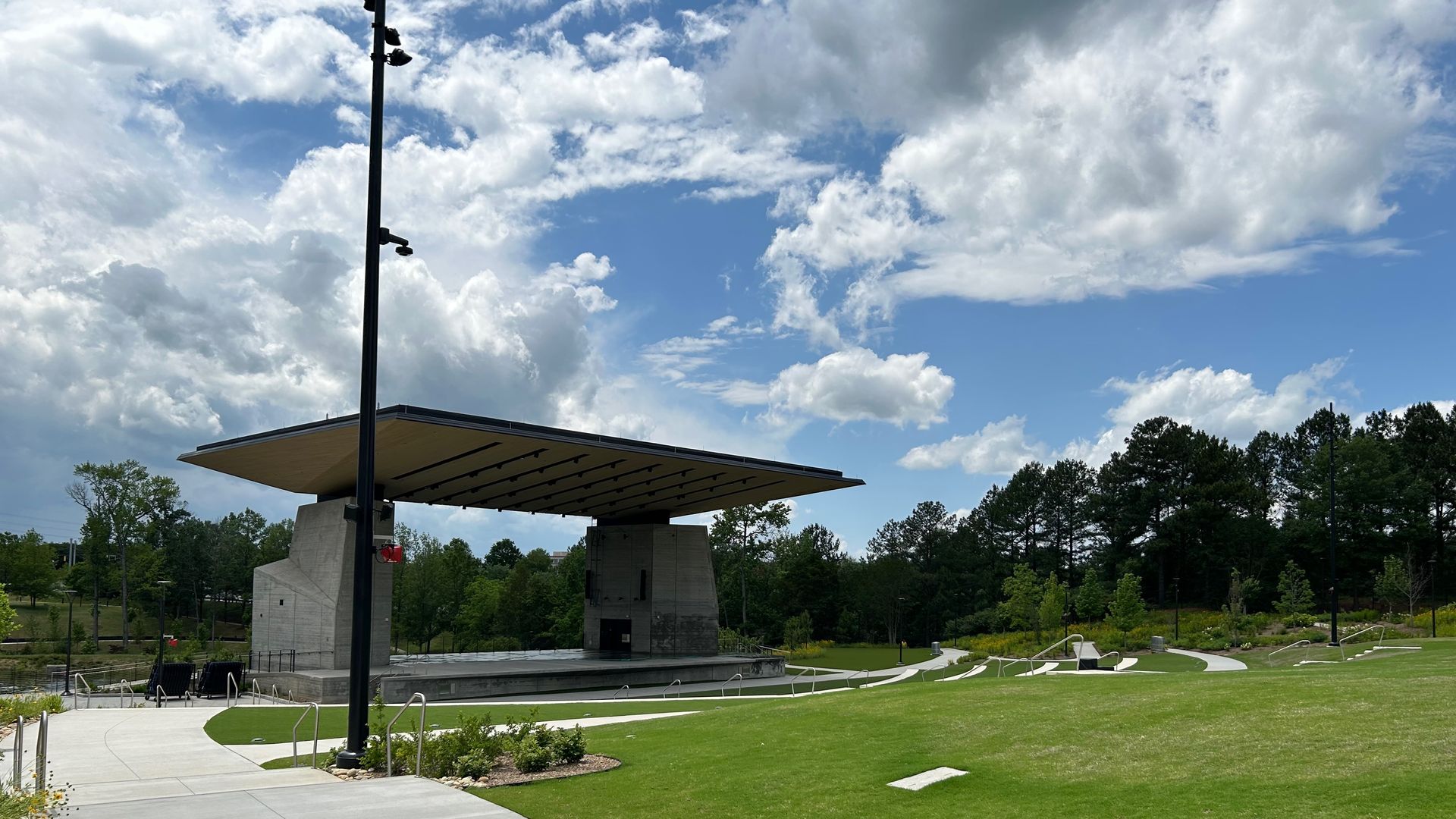 Outdoor amphitheater with a large concrete stage and black roof, surrounded by green grass, paved walkways, trees, and a partly cloudy blue sky.