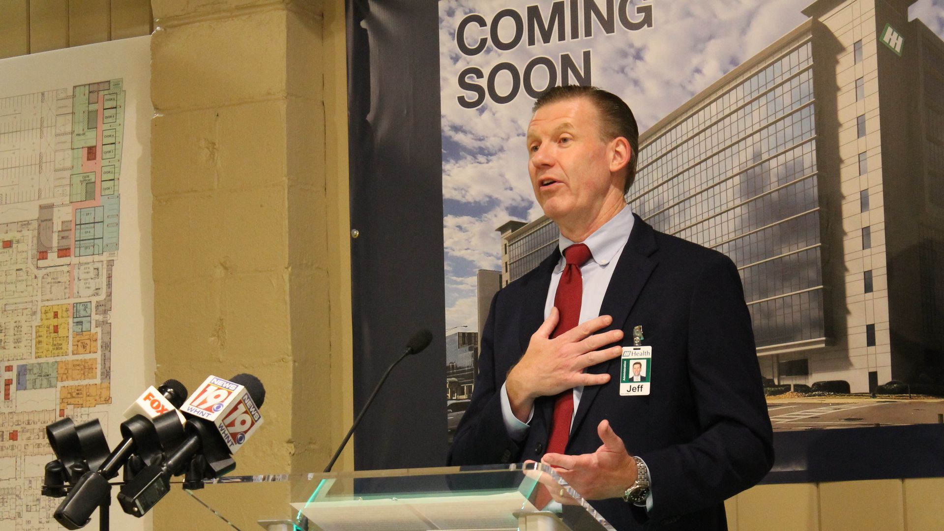 A man in a dark suit and red tie speaks at a glass podium with multiple microphones. Behind him, a banner shows a tall glass building and reads "COMING SOON."