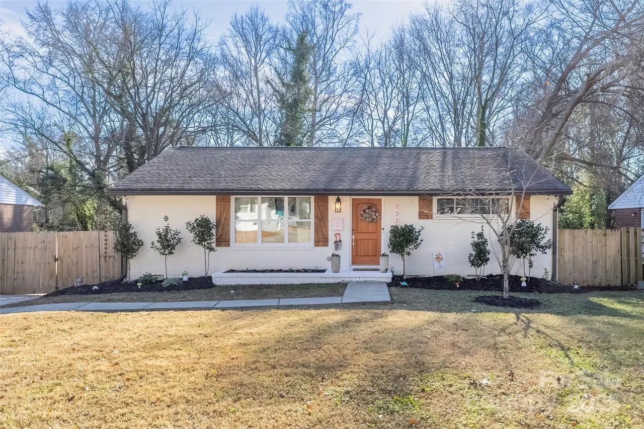 Single-story white house with brown roof and door, wooden shutters on windows, small garden with bushes, and leafless trees in background under a clear sky.