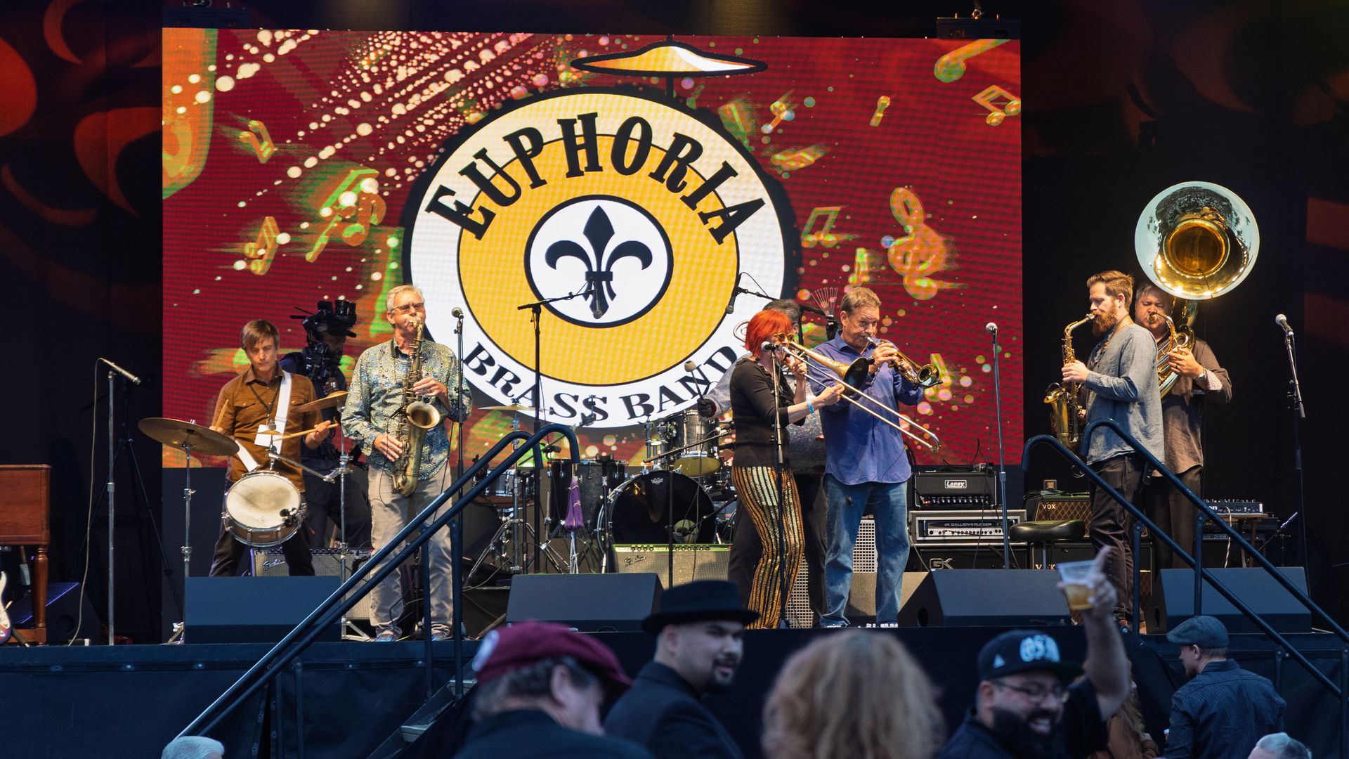 Brass band performs on stage; a large circular logo behind reads "EUPHORIA BRASS BAND" with colorful confetti on a red backdrop; audience visible in foreground.