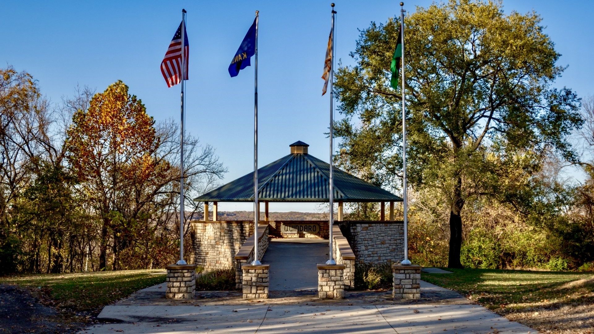 A park pavilion with a green metal roof and stone walls stands center, flanked by four flagpoles with flags; autumn trees and a clear blue sky surround.