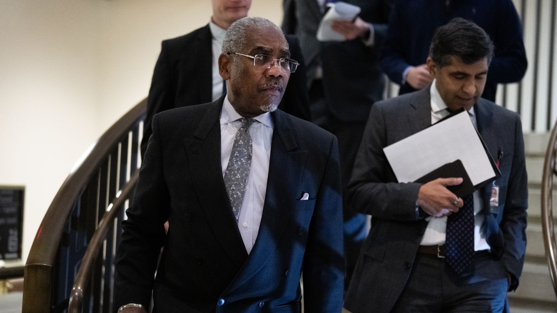 Group of men in business attire walking down stairs in an indoor setting, one man in the foreground wears a dark suit and patterned tie, another holds a folder with papers.