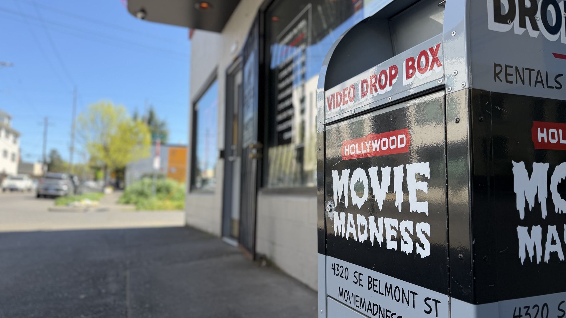 Close-up of a metallic video drop box outside a storefront on a sunny street. It shows bold white "MOVIE MADNESS" with a red "HOLLYWOOD" banner and "VIDEO DROP BOX" label.