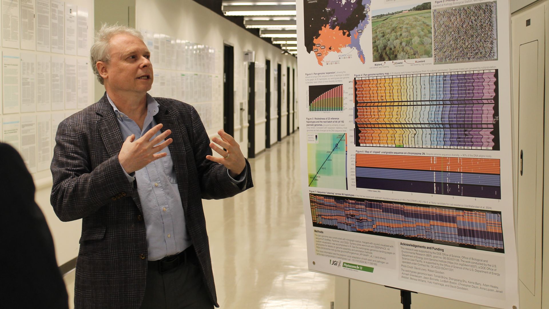 A man in a blazer explains a large scientific poster on a display board in a hallway lined with papers; the board features a map, colorful charts, and dense text, with a green EXIT sign overhead.