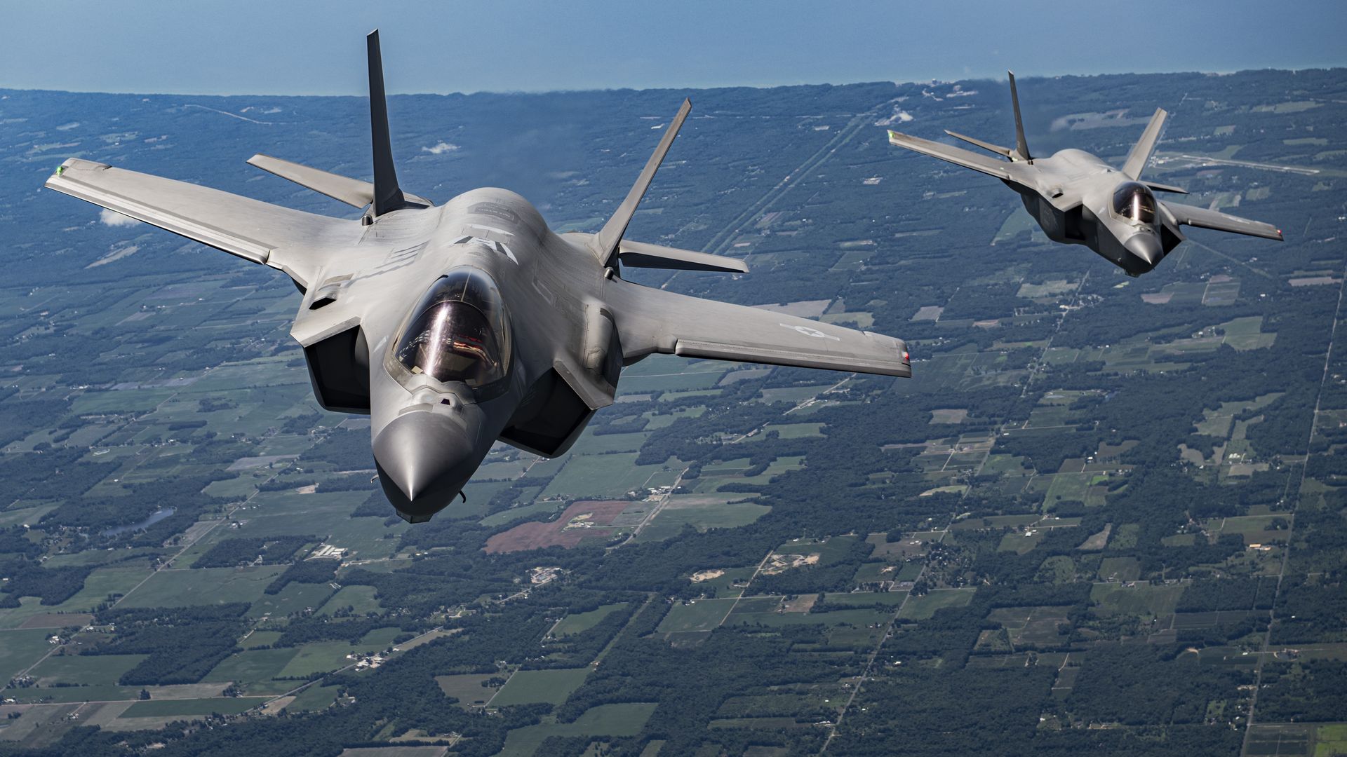Two gray fighter jets flying over a green and brown patchwork of farmland and forest under a clear blue sky.