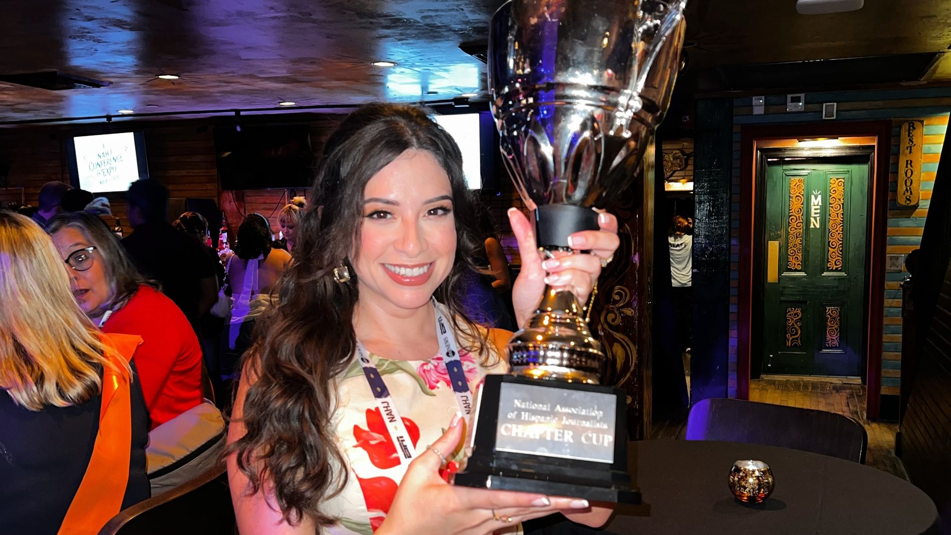 Smiling woman in a white floral dress holds a large silver trophy with a black base at a crowded, dim venue with colorful lighting and people in the background.