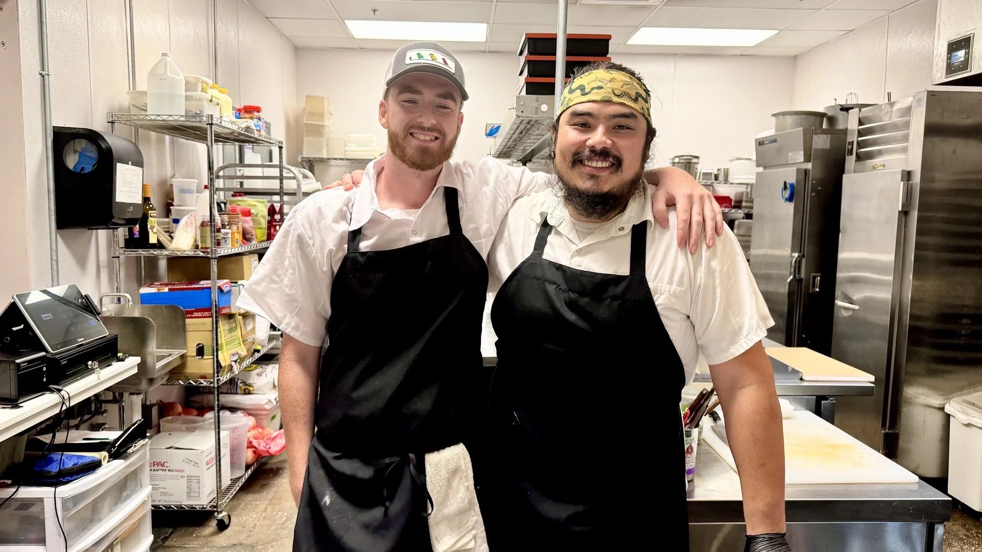 Two smiling chefs in white shirts and black aprons standing in a commercial kitchen with metal shelves, refrigerators, and cooking equipment.