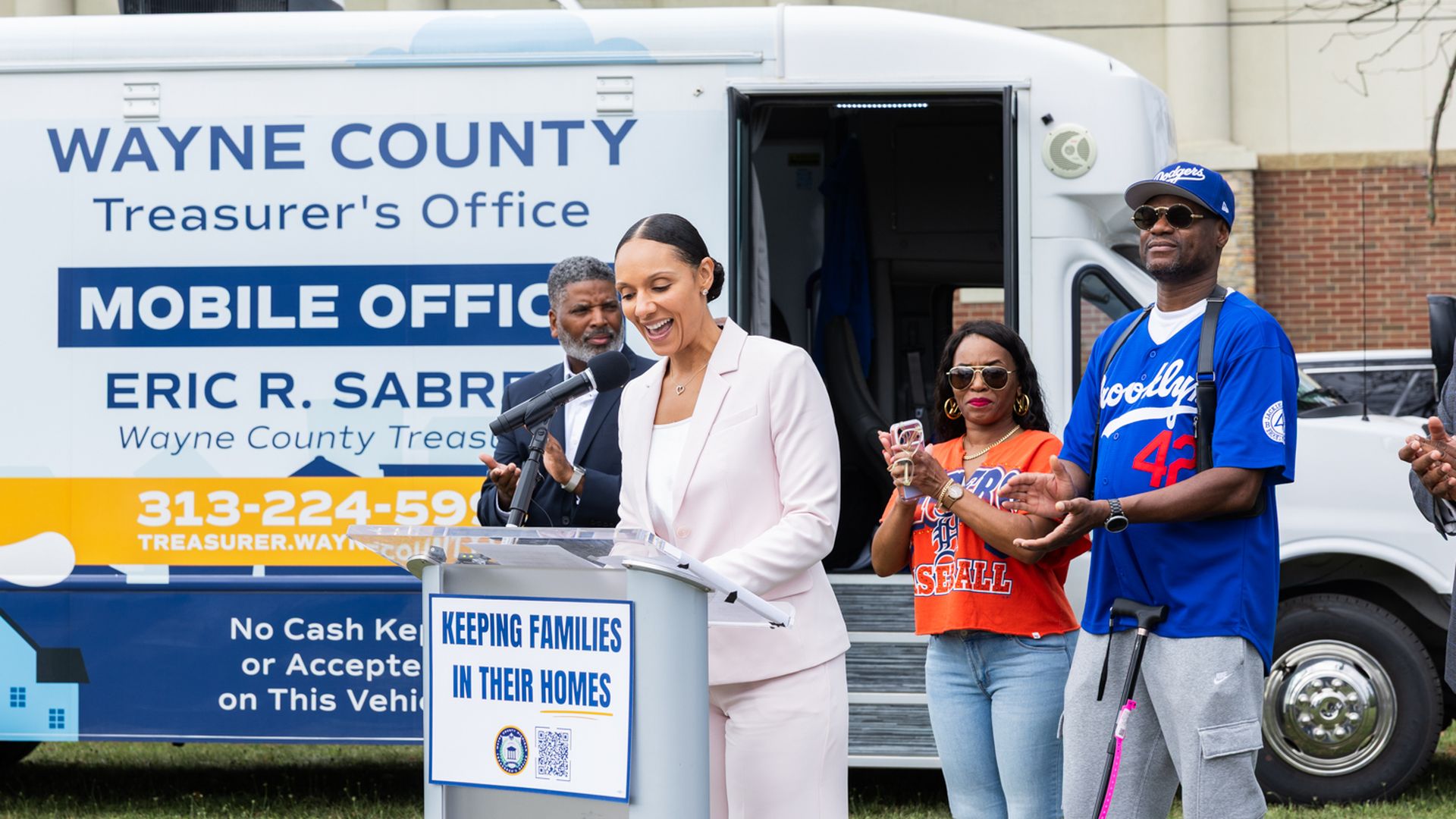 Speaker in a pale pink suit at a podium beside the Wayne County Treasurer's Office Mobile Office van; lectern sign reads 'KEEPING FAMILIES IN THEIR HOMES' as supporters stand nearby.