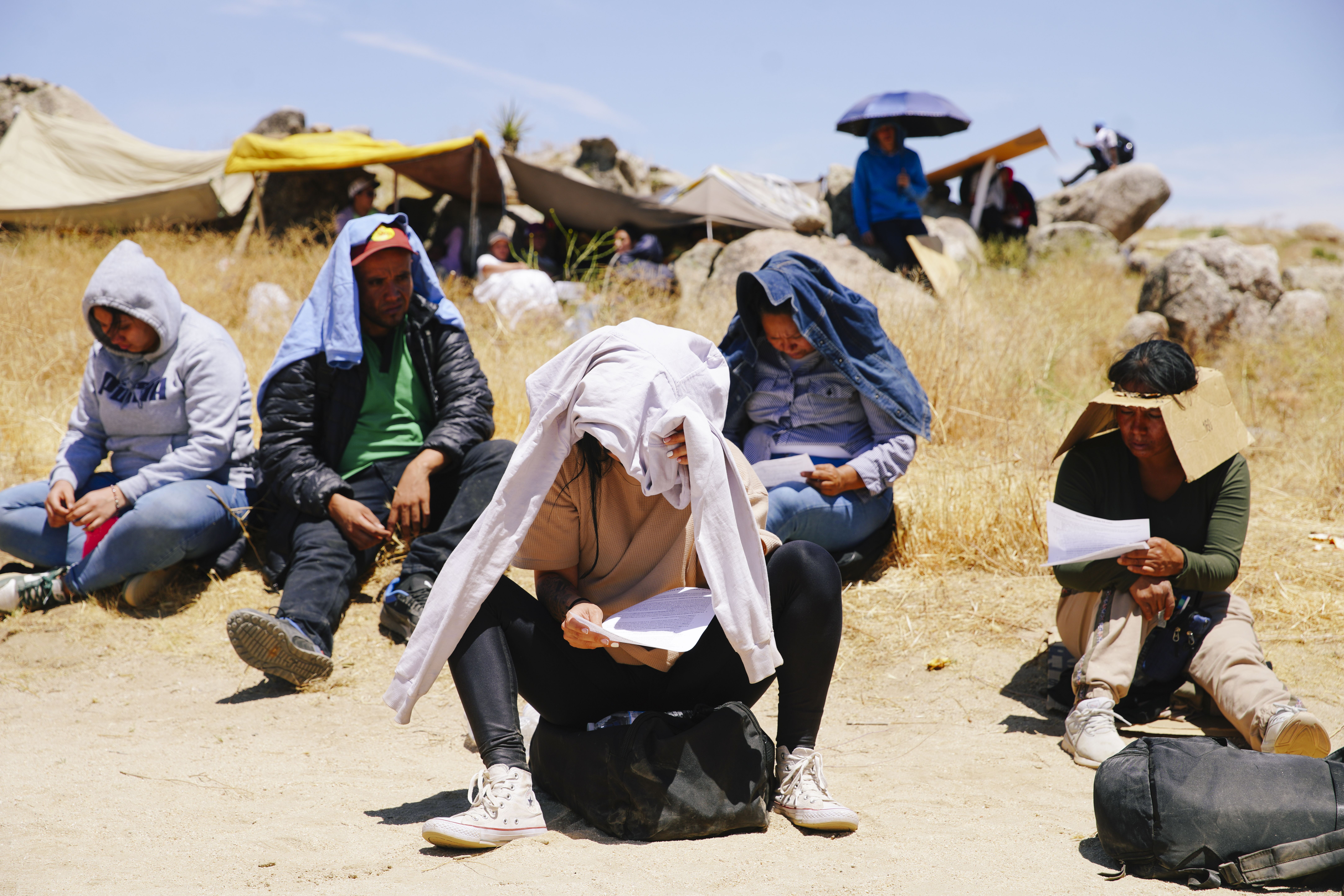 A group of migrants sitting on the ground, each one has covered their face and neck with a shirt to protect themselves from the sun. 