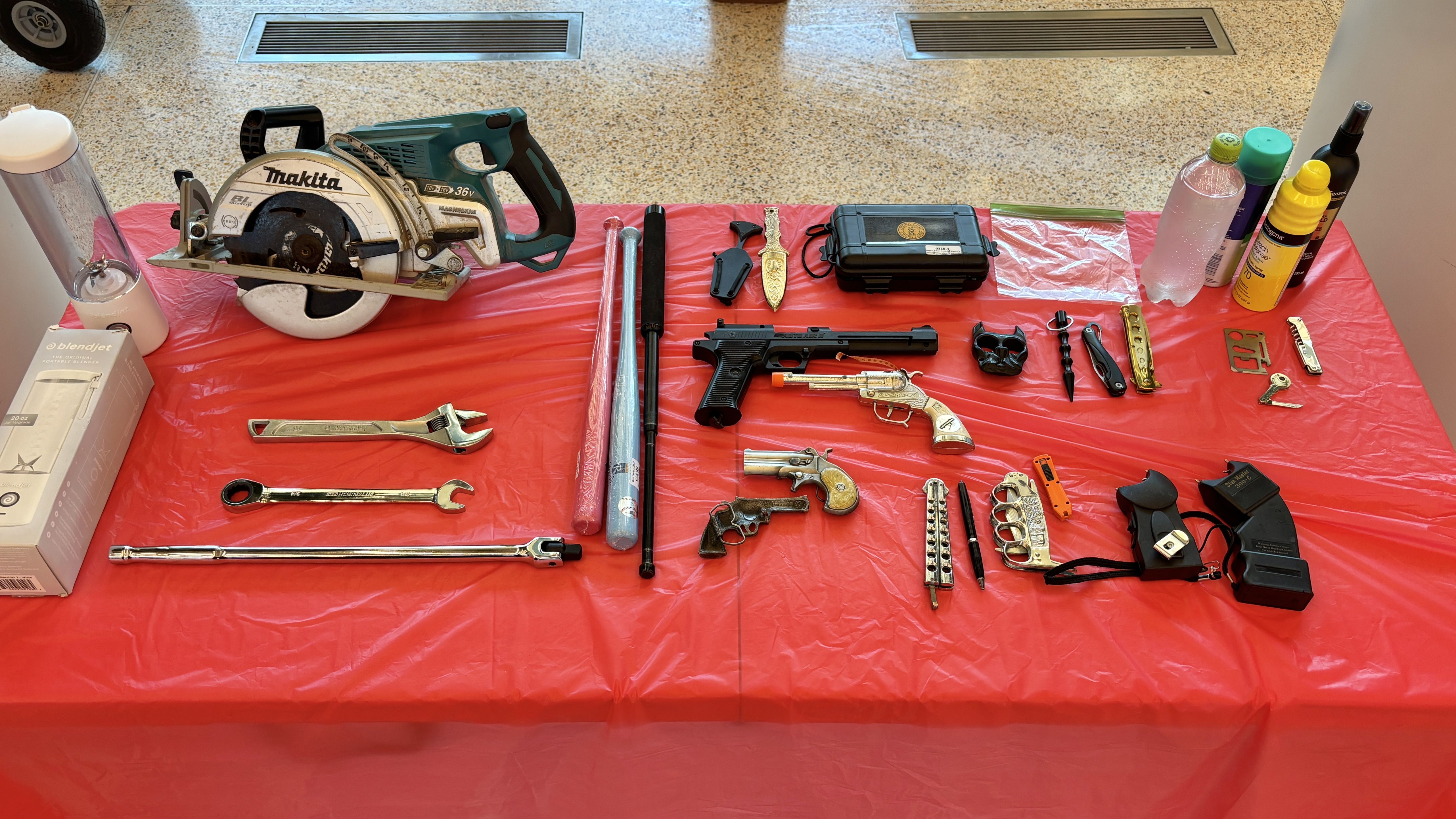 On a red tablecloth lies an array of tools and weapons: a Makita circular saw, a green cordless drill, a black pistol, an ornate gold pistol, knives, wrenches, a black case, and cleaning bottles to the right.