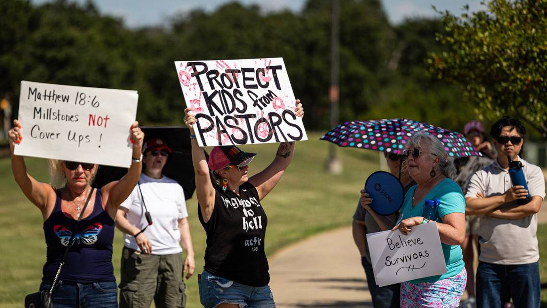 Protesters hold up signs saying "Matthew 18:6 Millstones not cover ups," "Protect kids from pastors" and "Believe survivors," near grass and vegetation outside Gateway Church in Southlake, Texas, protesting  following child sexual abuse allegations against Robert Morris.