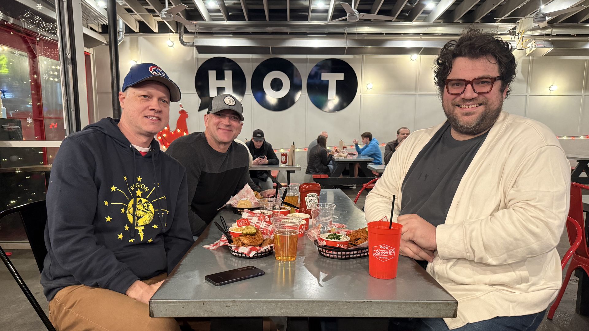Three men smiling at a metal table in a casual restaurant with red chairs, food baskets, drinks, and a wall sign reading "HOT" behind them.