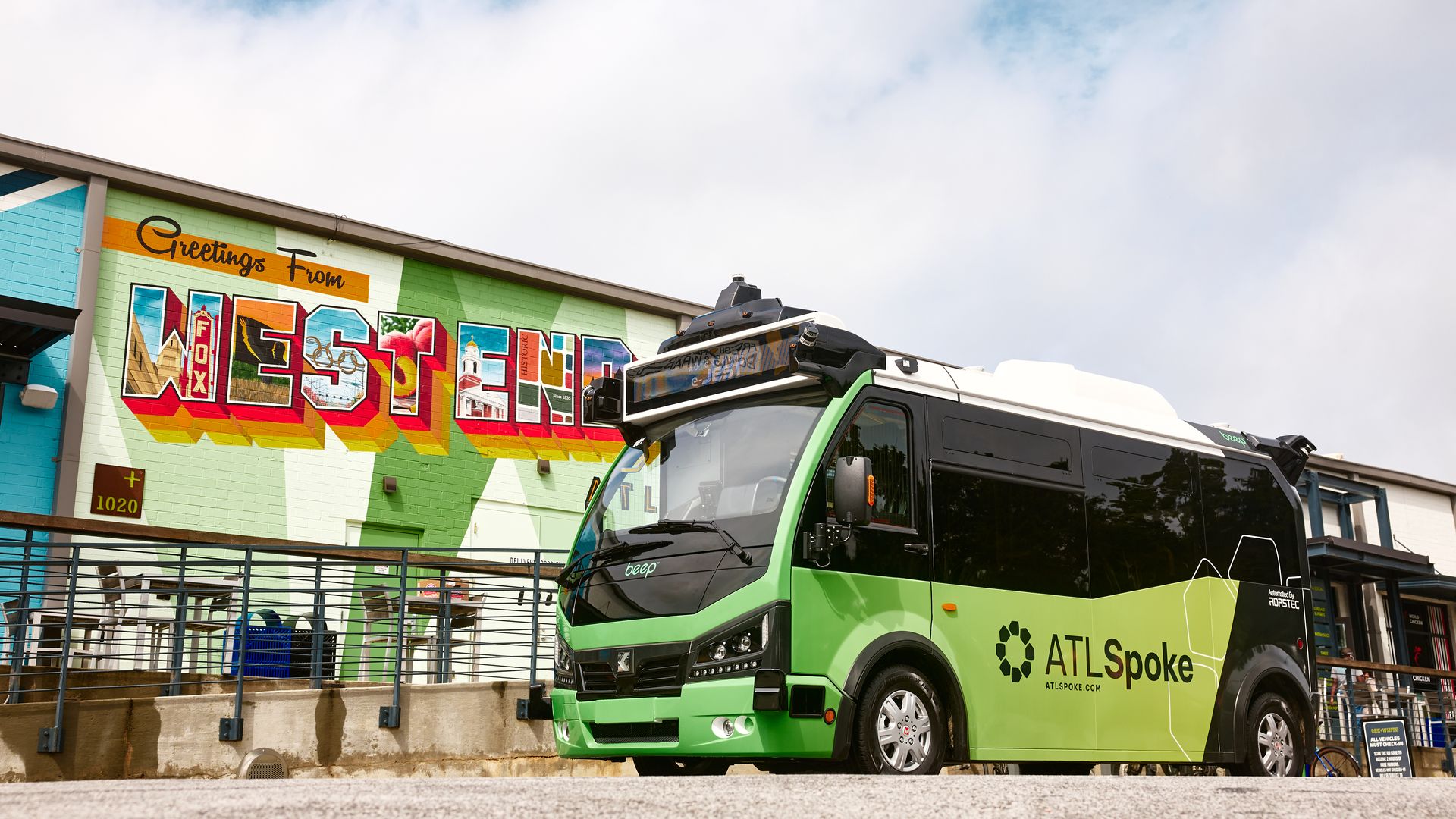 Colorful mural on a building reads "Greetings From" with large, vibrant "WEST" letters; a lime-green ATL Spoke bus sits in front under a blue sky with white clouds.