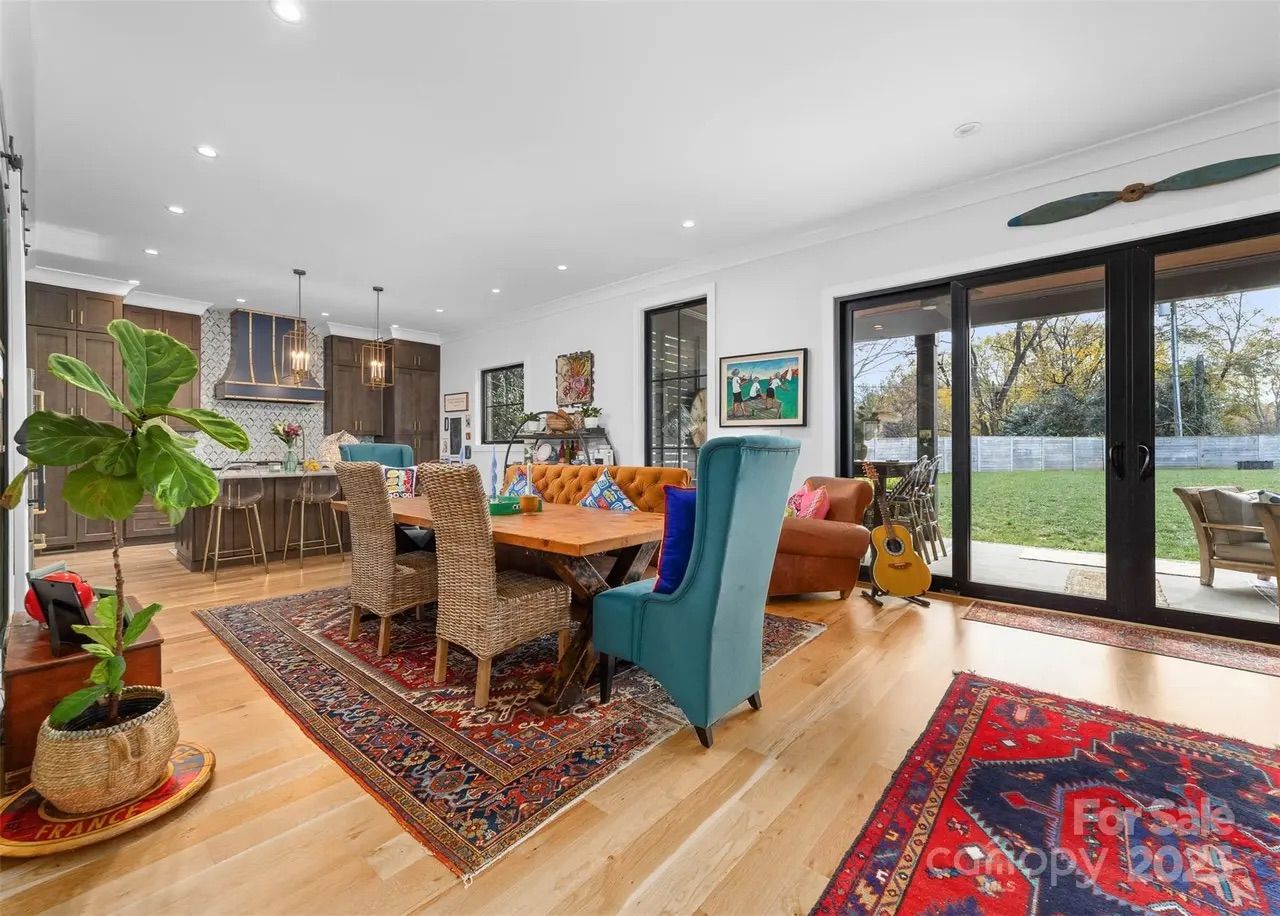 Bright open-concept living space with hardwood floors, colorful patterned rugs, a wooden dining table with wicker chairs and teal wingback chair, orange couch, large sliding glass doors, and a guitar.