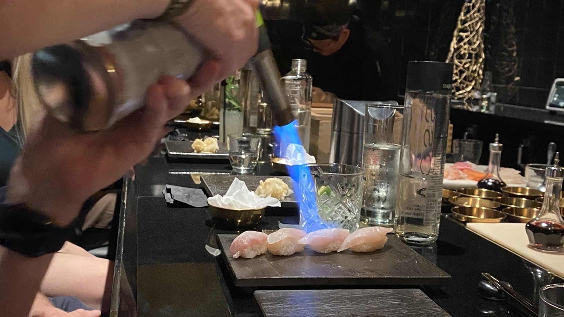 A person fires a flaming blowtorch at four pieces of sushi on a black plate. 
