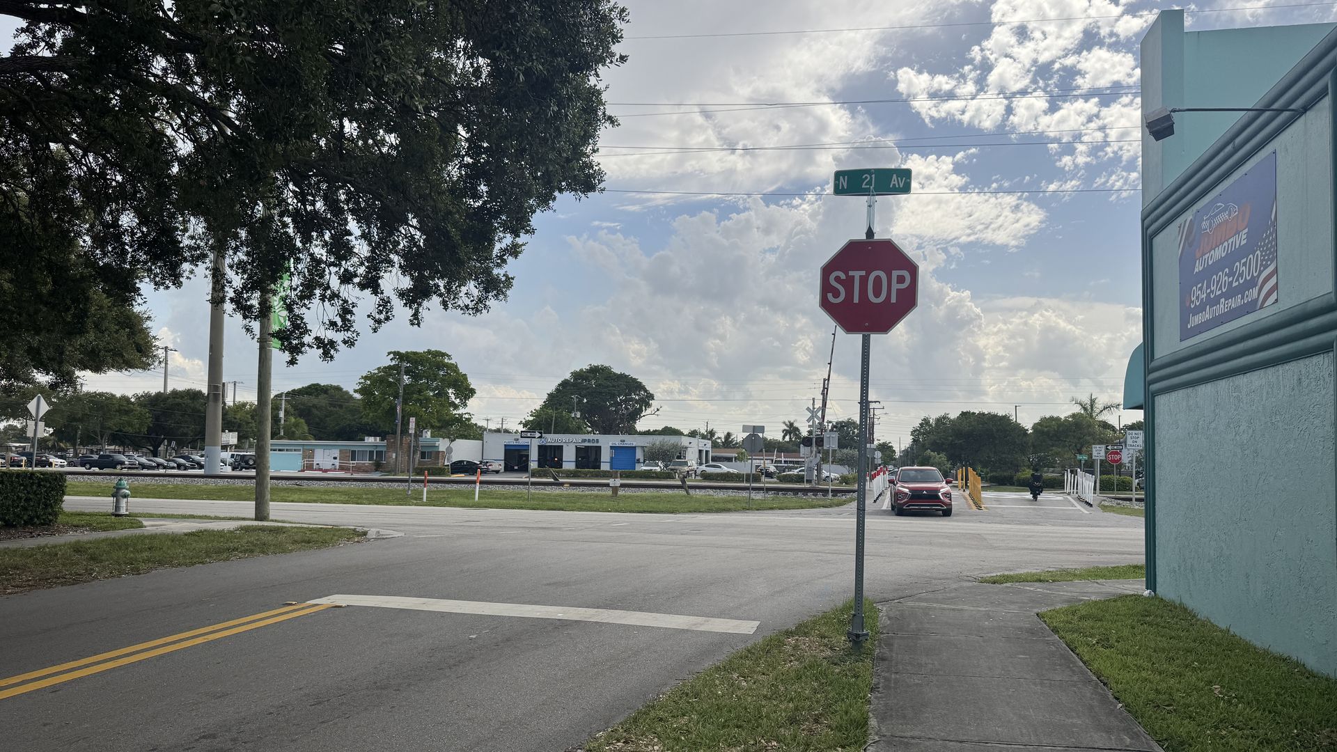 Wide street intersection under a blue sky with clouds. A red STOP sign and green street sign reading N 21 Av stand beside pale blue building. Left tree dominates; distant cars and storefront visible.
