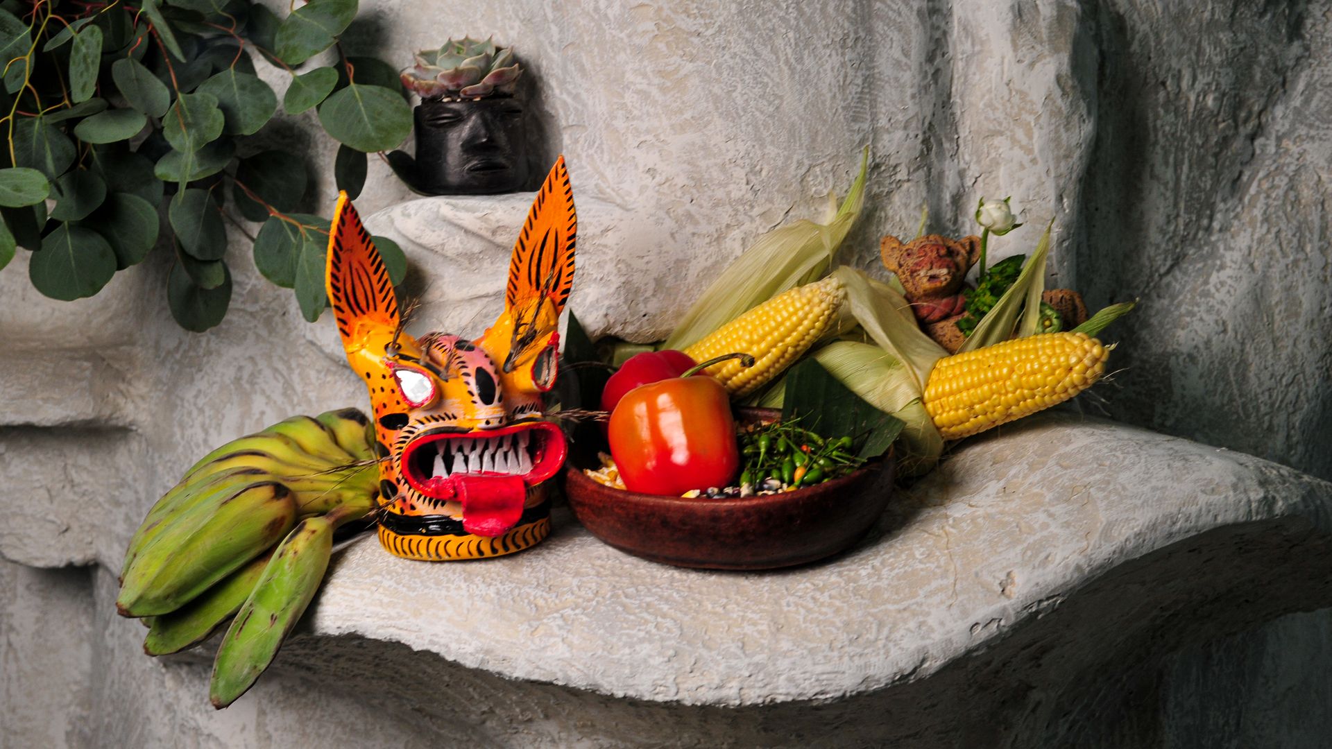 Decorative altar on a textured stone ledge with a bright orange tiger mask, a black face pot crowned by succulents, and corn, peppers, and green eucalyptus leaves.