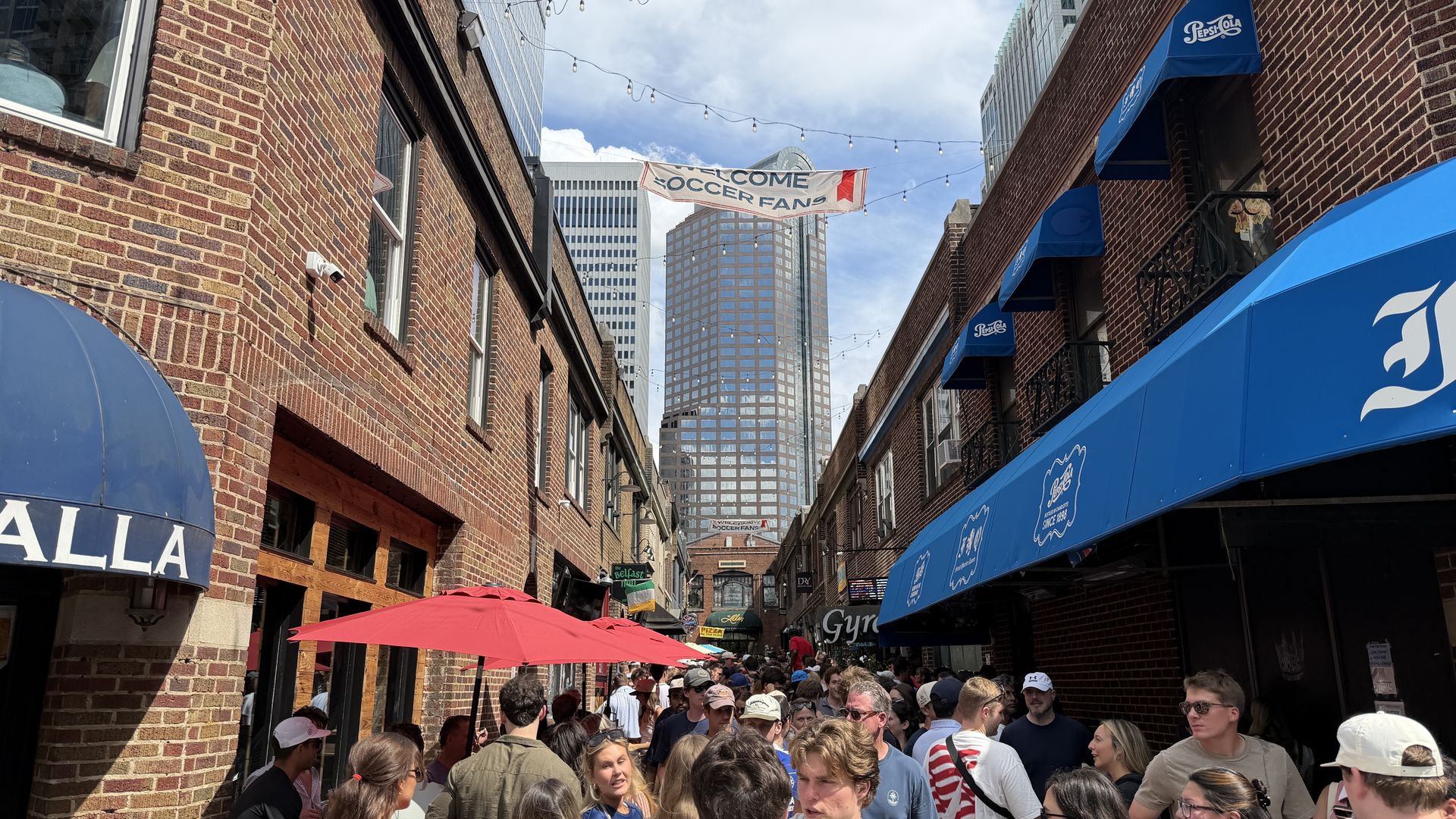 Busy alley between brick buildings with blue awnings and string lights; a banner reading Welcome Soccer Fans spans the street as a crowd gathers under red umbrellas and glass buildings loom.