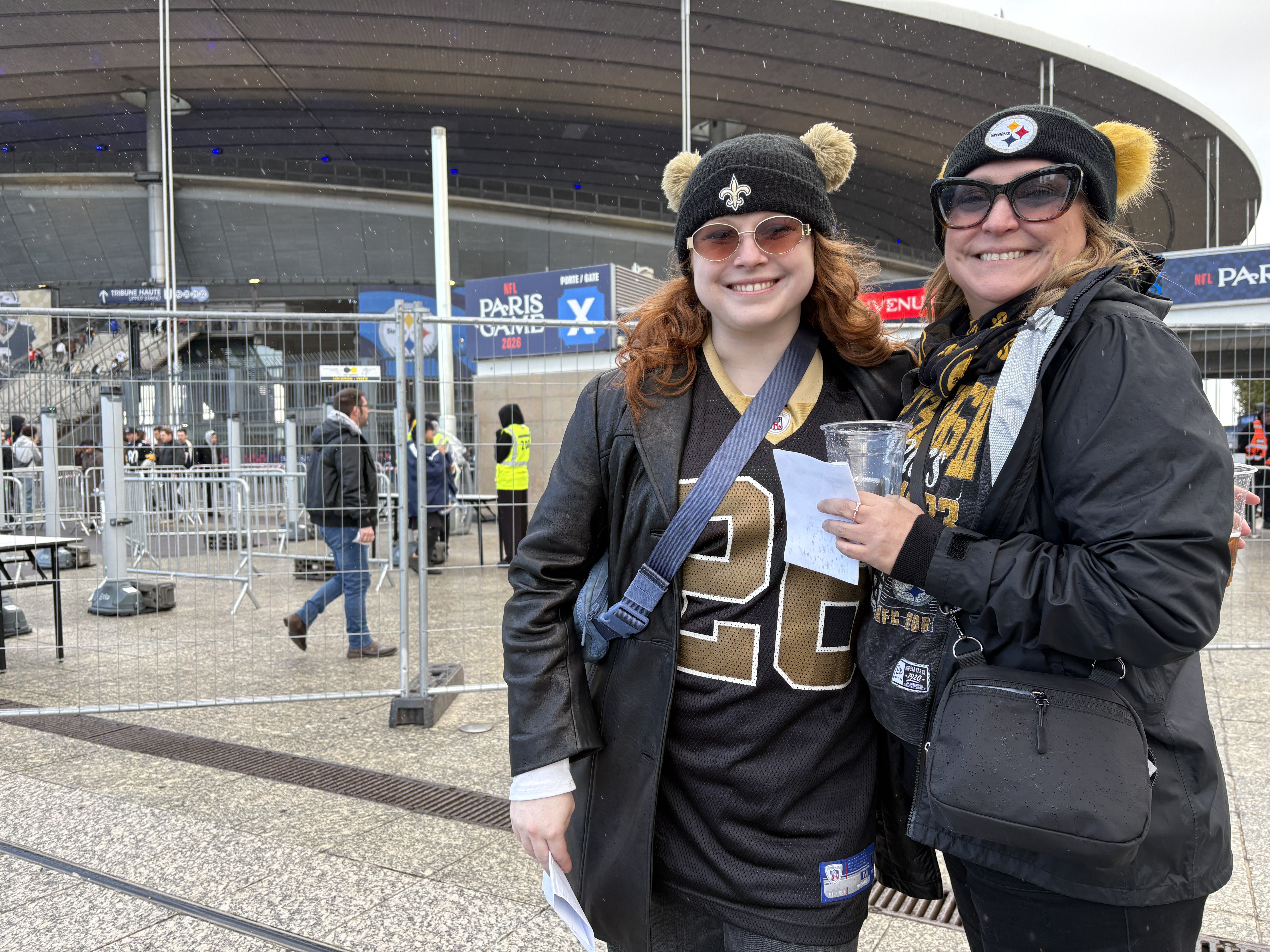 Two smiling fans pose outside a stadium with metal barriers in falling snow. Left: black beanie with a fleur-de-lis and bear ears, sunglasses; right: Steelers beanie with ears and large sunglasses.