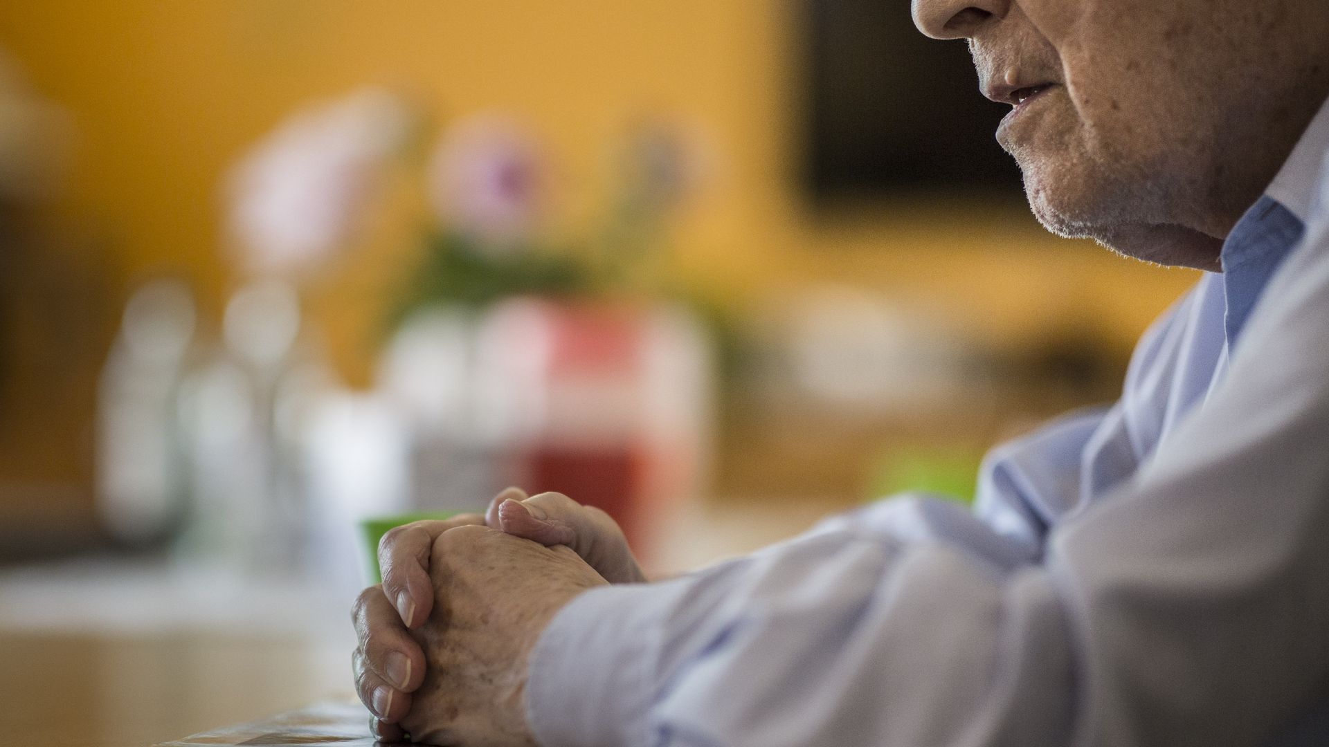 An elderly man sits at a table with his hands folded.