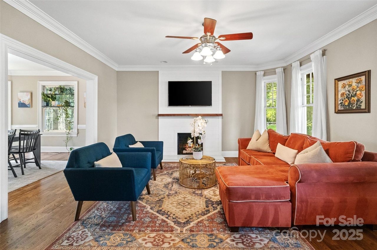 Living room with a rust-colored sectional sofa, two dark blue armchairs, a patterned rug, a white fireplace with TV above, and large windows with sheer white curtains.