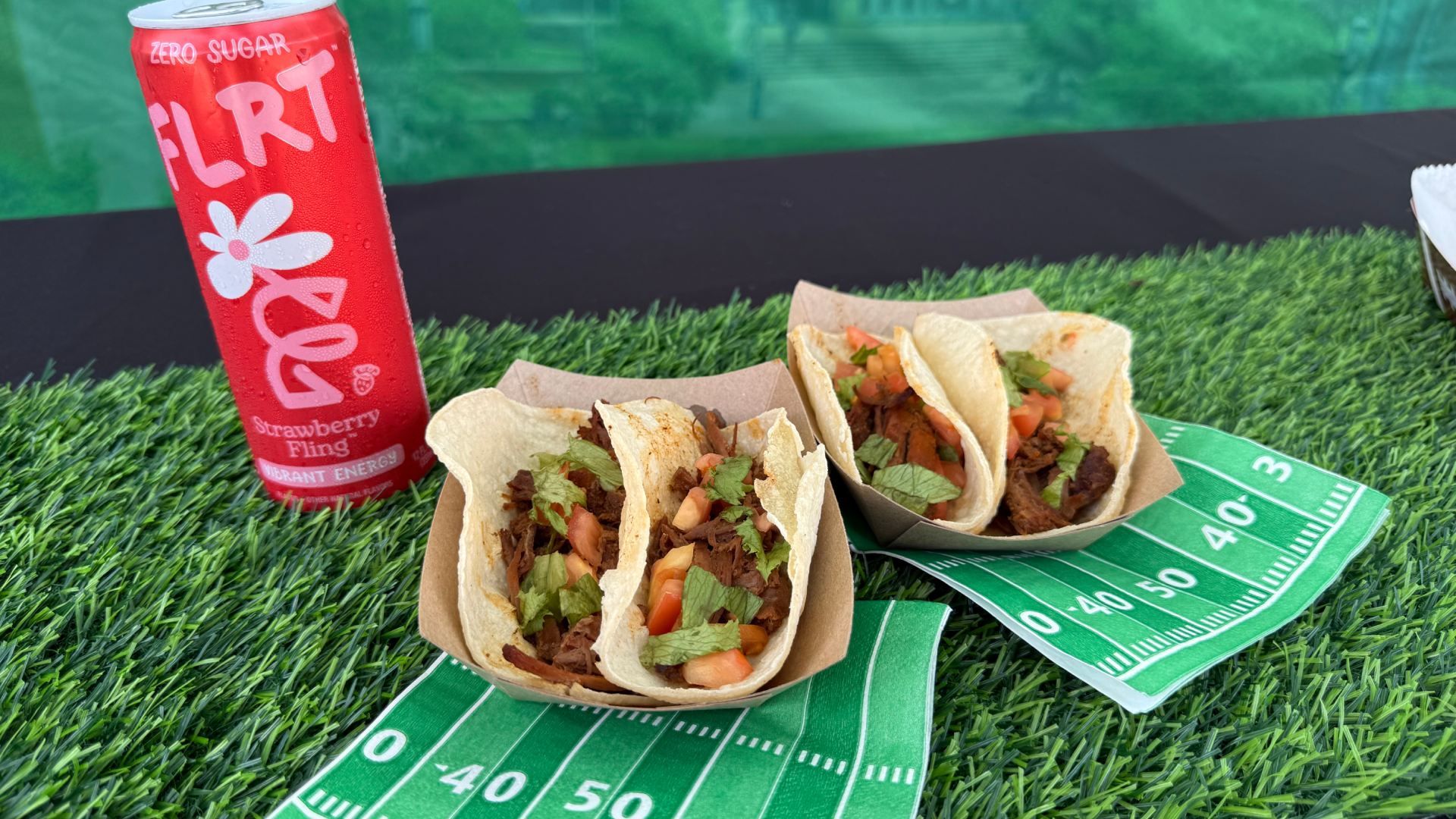 A red can of Flirt Zero Sugar labeled "Strawberry Fling" sits on artificial grass beside three beef tacos filled with cilantro and diced tomatoes on a green yardage‑measuring mat.