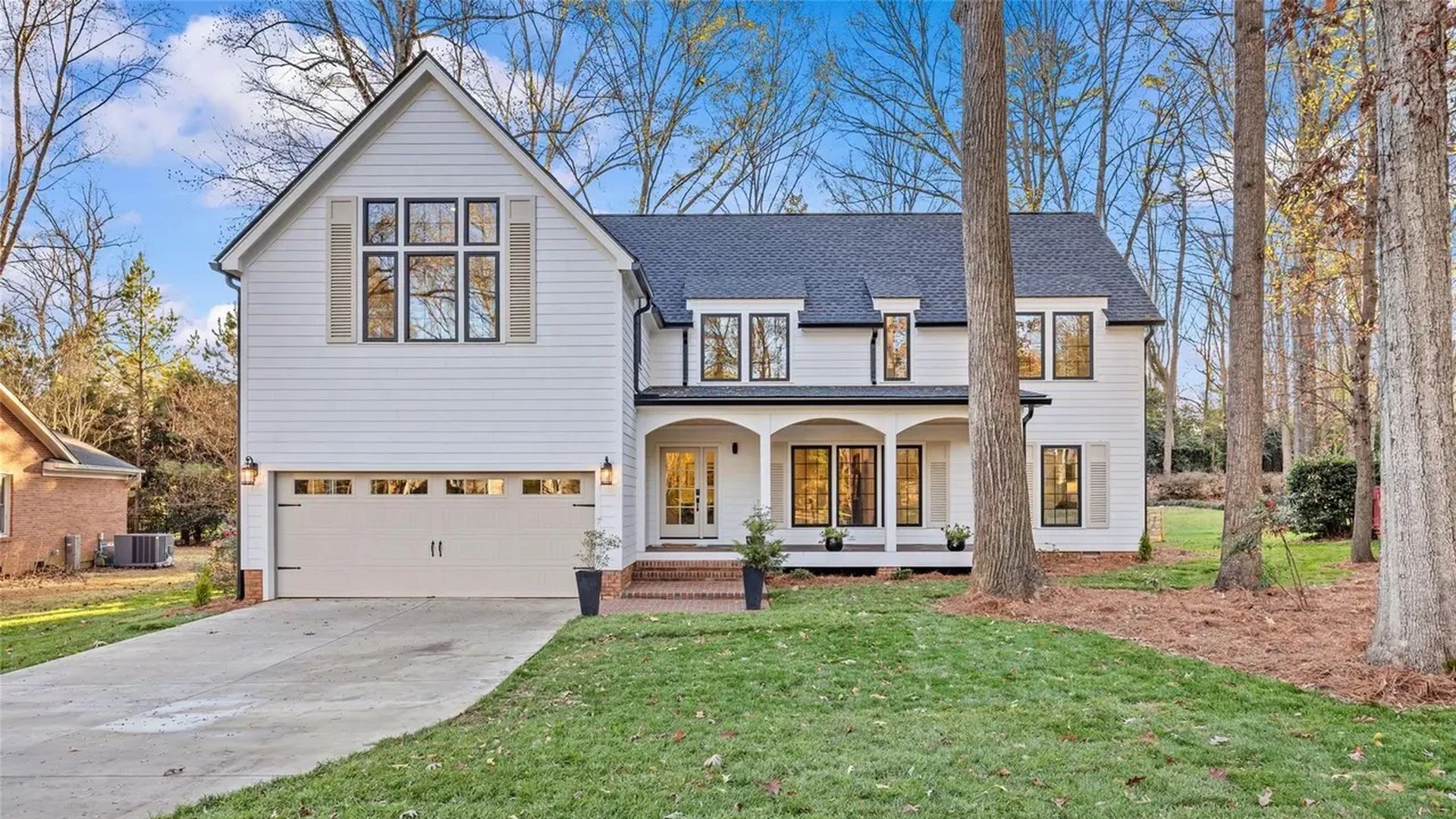 White two-story house with black roof, large windows, front porch with three arches, two potted plants by entrance, large driveway, lawn, and tall leafless trees under blue sky.