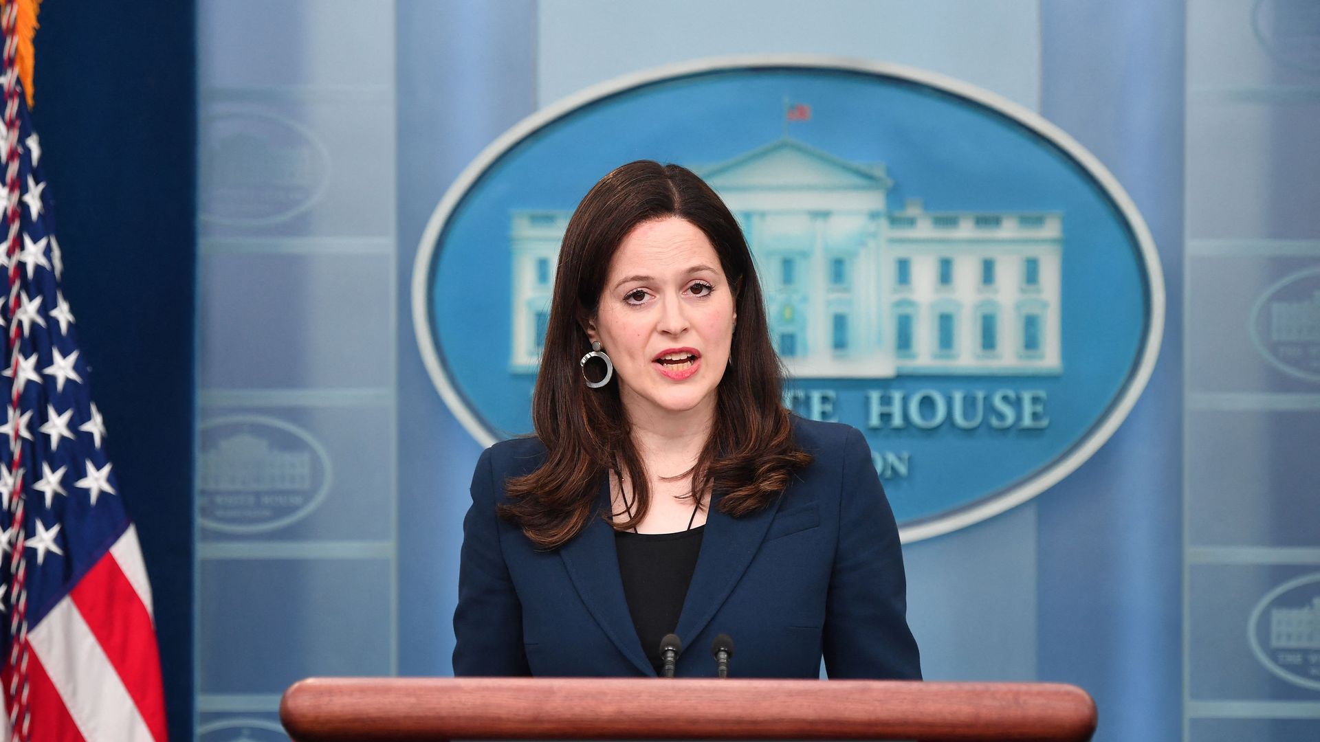 A woman in a dark blue blazer speaks at a podium labeled "The White House Washington" with the American flag and White House emblem in the background.