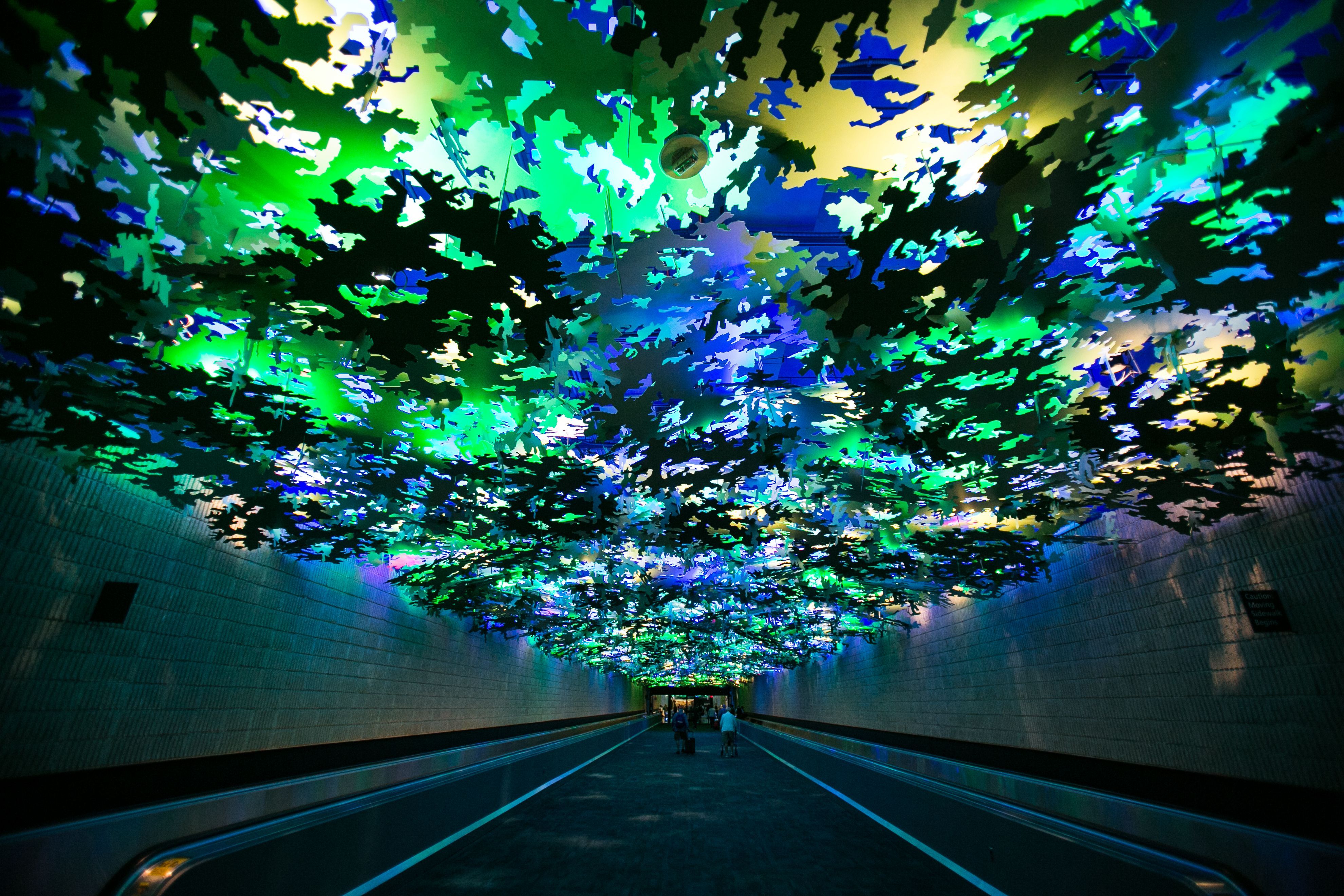 People walk in a long windowless corridor with moving walkways in an airport. A tree canopy-like artwork is suspended from the ceiling
