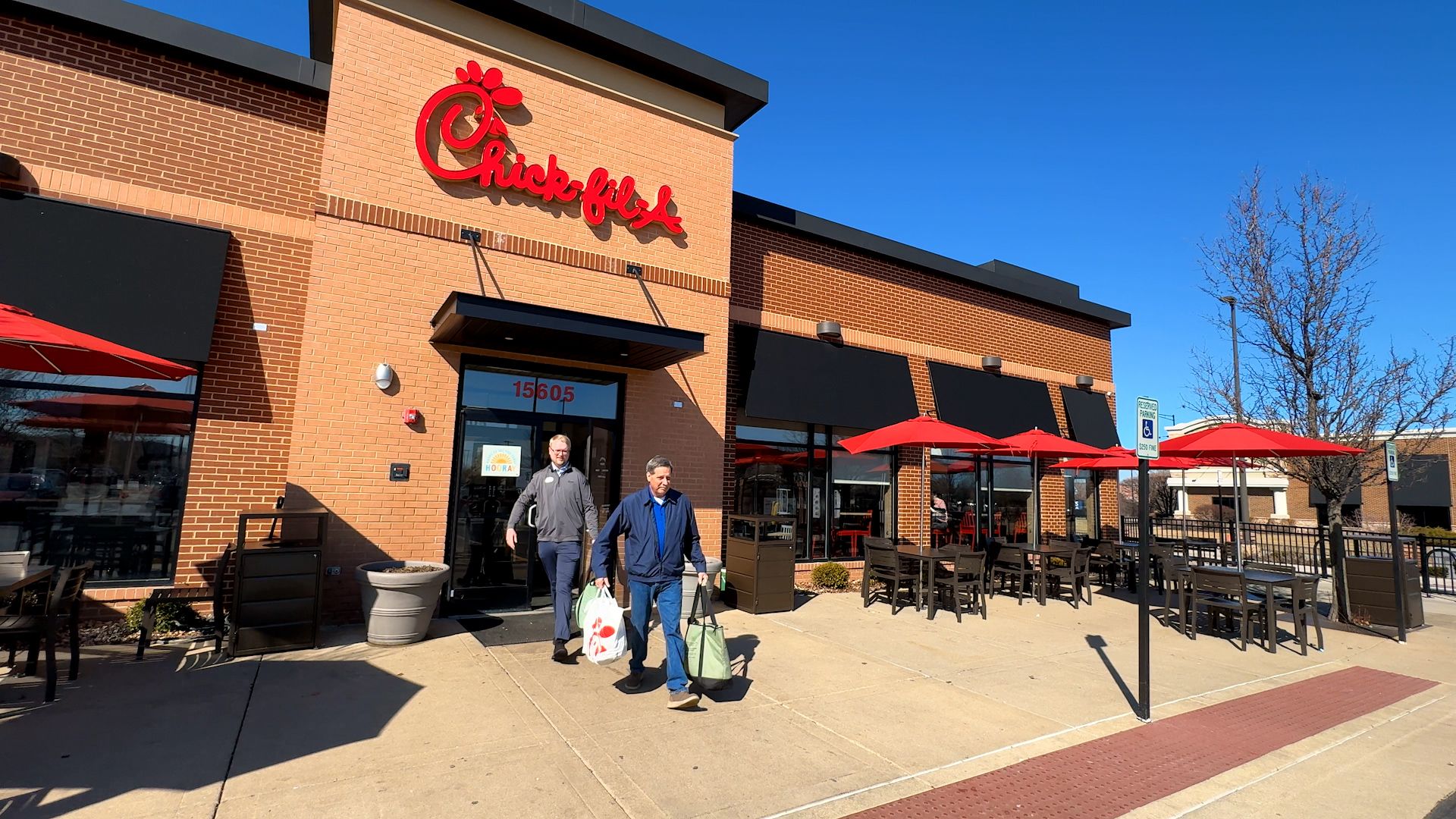 Two men carrying takeout bags walking out of a Chick-fil-A restaurant with brick exterior, black awnings, and outdoor seating with red umbrellas on a sunny day.
