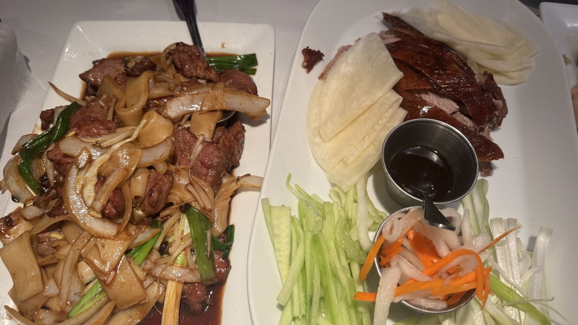 Two white plates on a table: left plate shows beef and onion stir-fry with wide noodles; right plate features sliced roast duck with thin pancakes, hoisin sauce, and cucumber–carrot salad.
