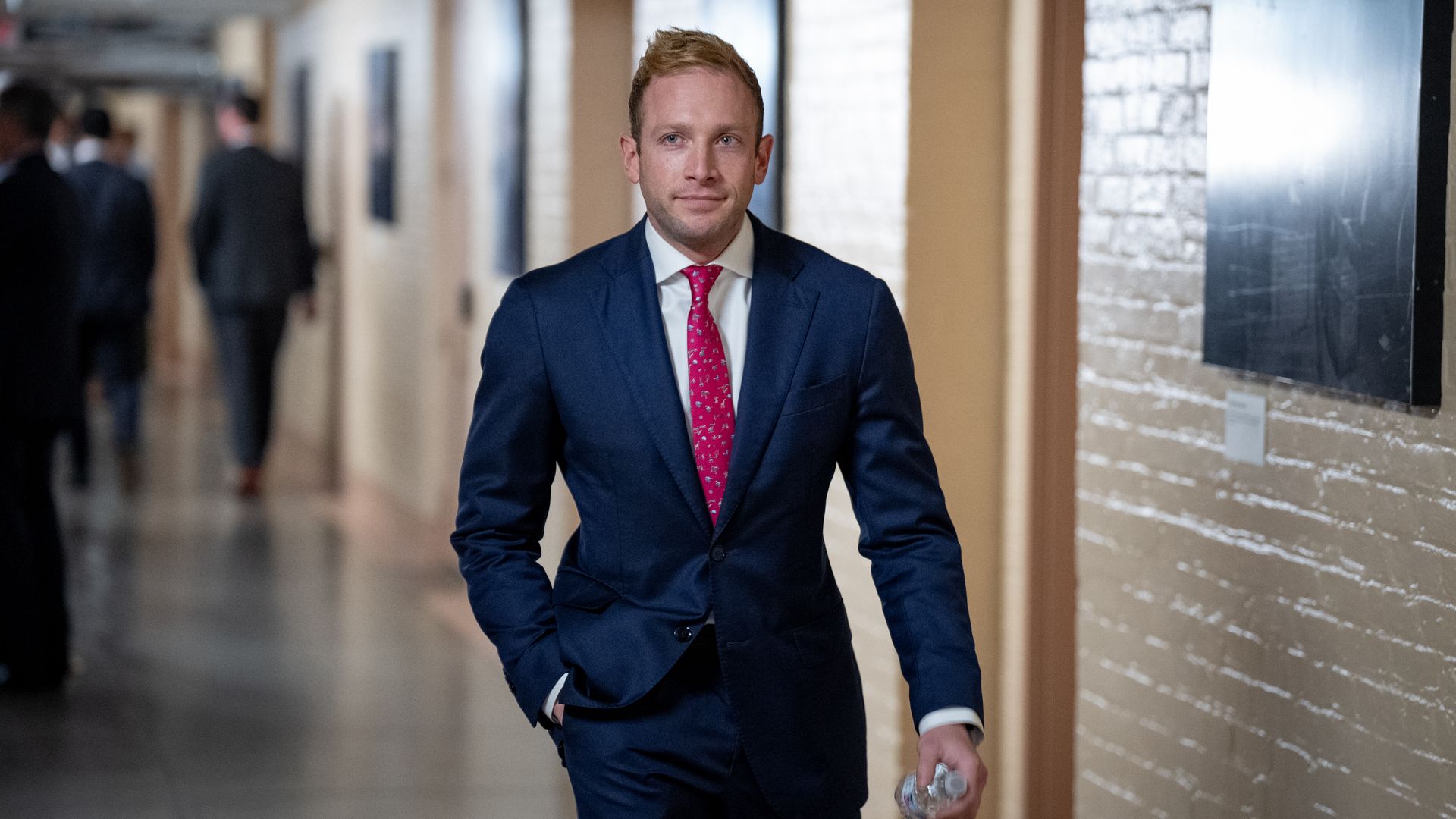 Rep. Max Miller, wearing a blue suit, walking through a white brick hallway while holding a water bottle.