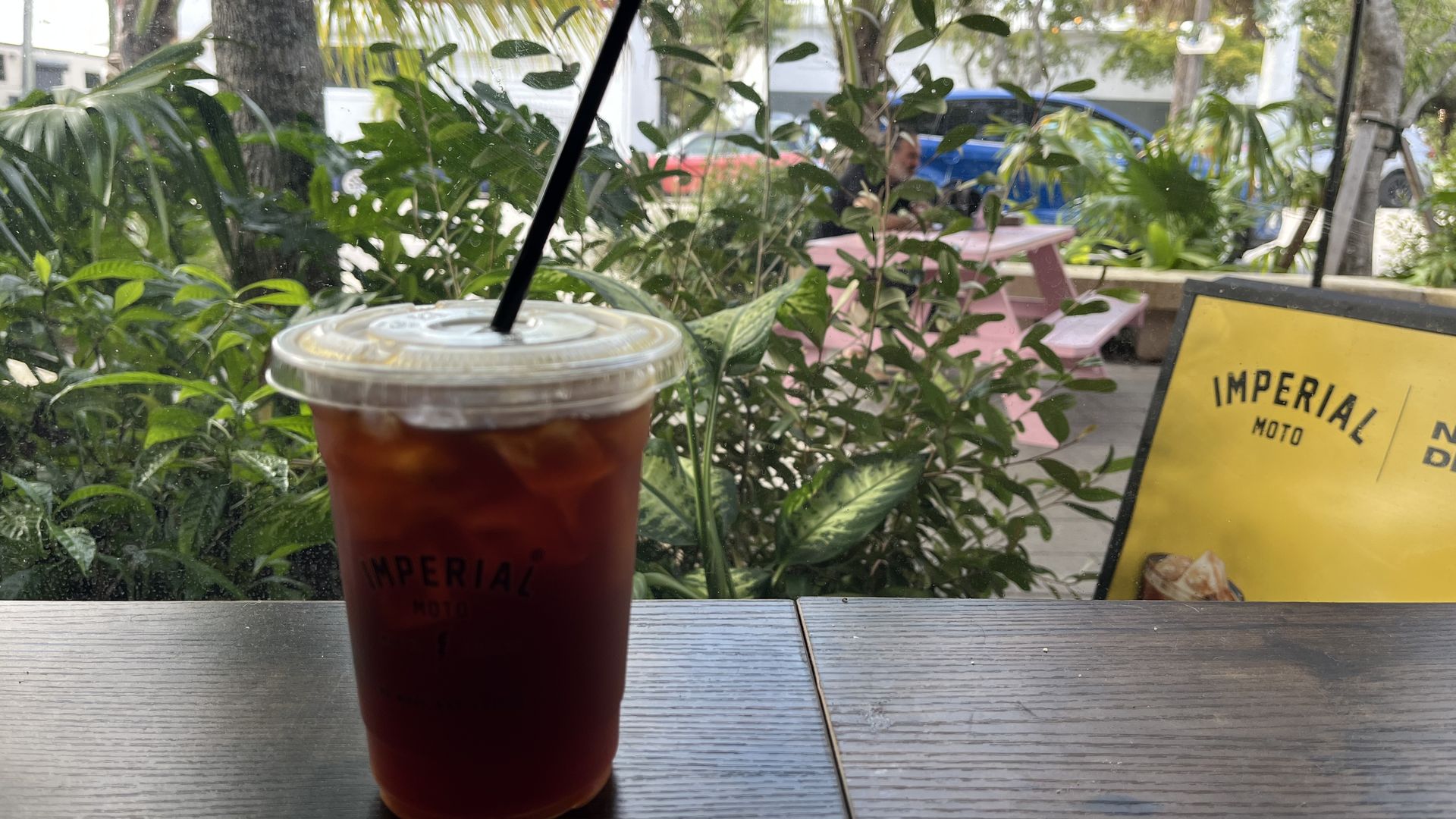 Iced coffee in a clear plastic cup with a black straw sits on a wooden table by a window. Green plants and outdoor seating, including pink benches and a yellow sign with "IMPERIAL MOTO" text, visible outside.