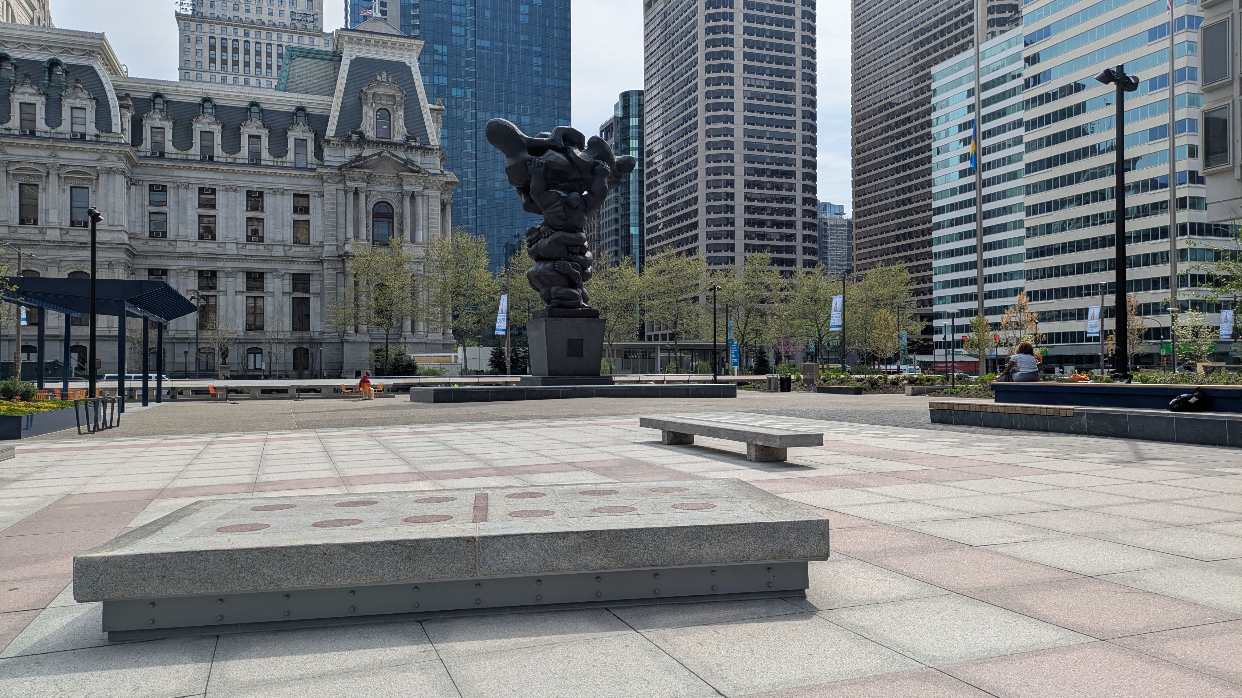 Wide city plaza with a large dark abstract sculpture on a pedestal in center, surrounded by tall glass towers and a historic stone building on the left. Benches, trees, and people sit around.