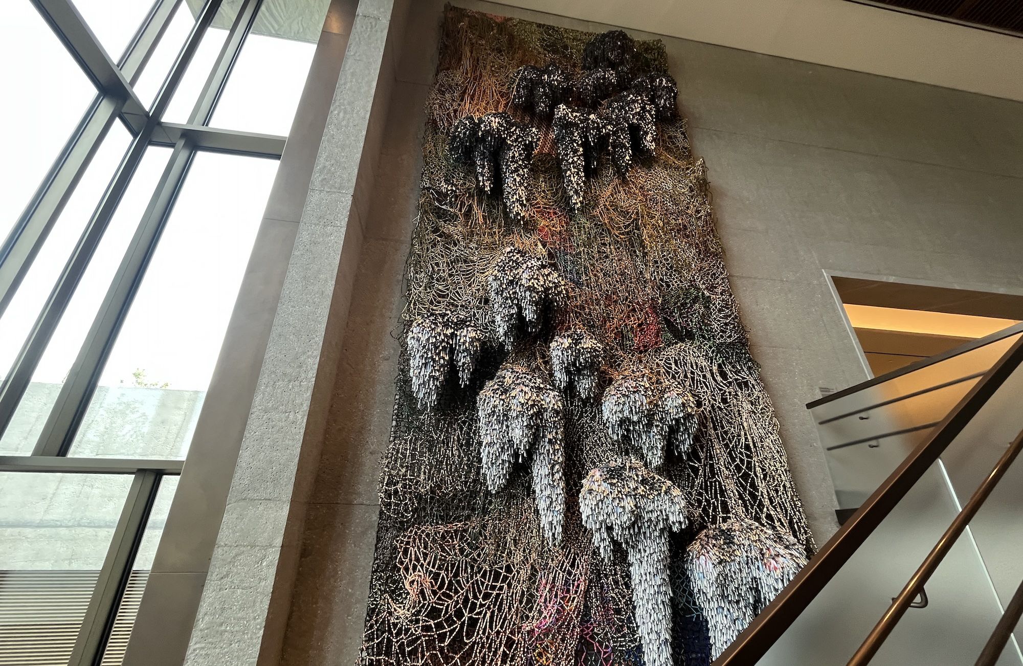Tall sculpture of interwoven metal mesh drapes down a wall beside a staircase in a modern atrium, featuring dark copper tones and white speckles against a gray interior.