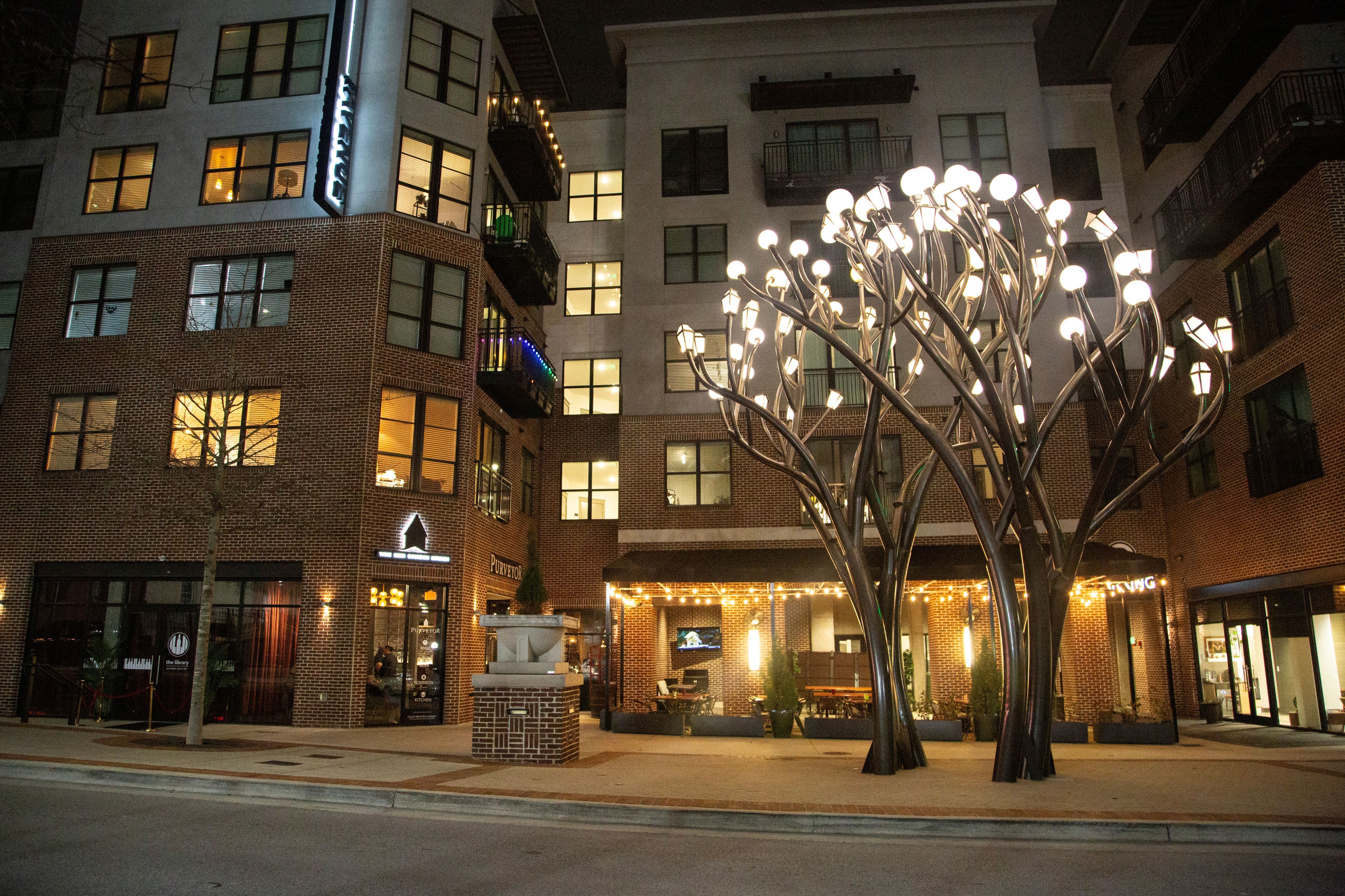 Night view of a modern apartment complex with brick and beige walls, featuring a large metal tree sculpture with multiple glowing light bulbs at the center of a courtyard.