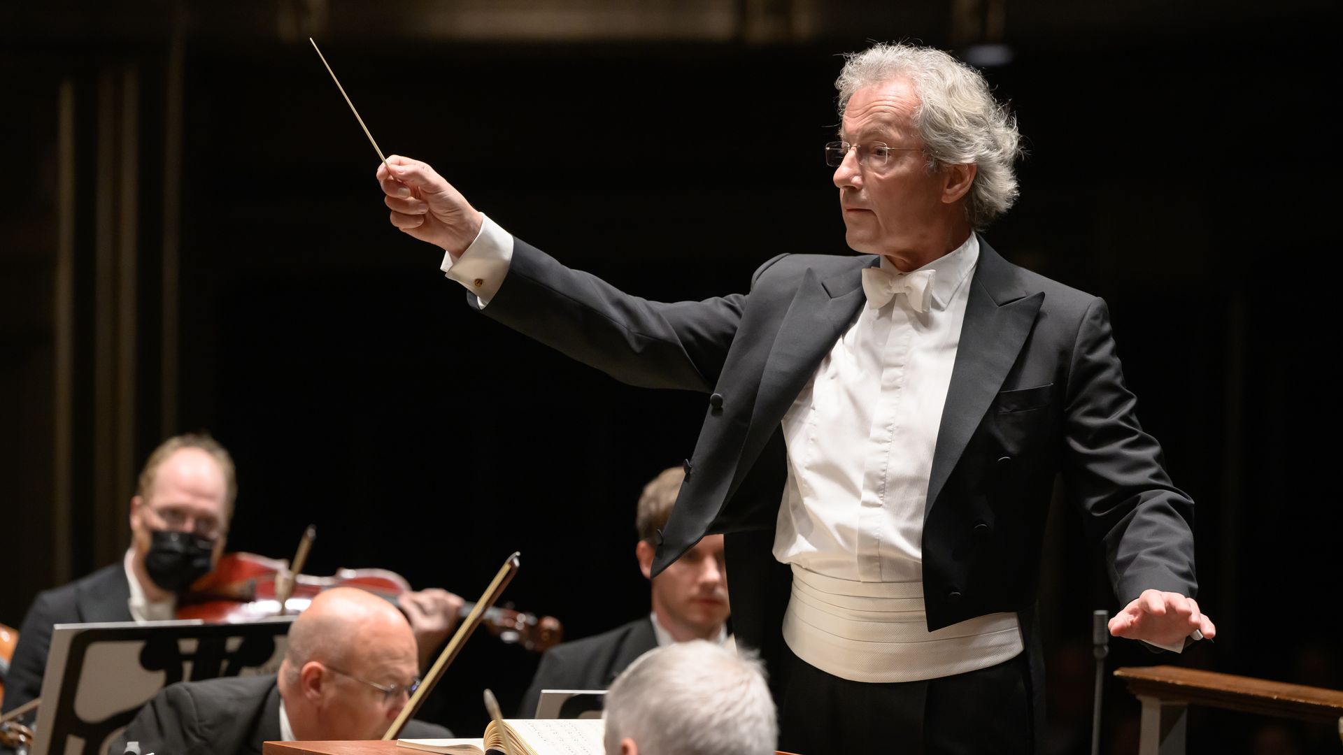Conductor in a black tuxedo and white bow tie raises a baton, directing an orchestra in a dark concert hall, with musicians and music stands visible behind him.