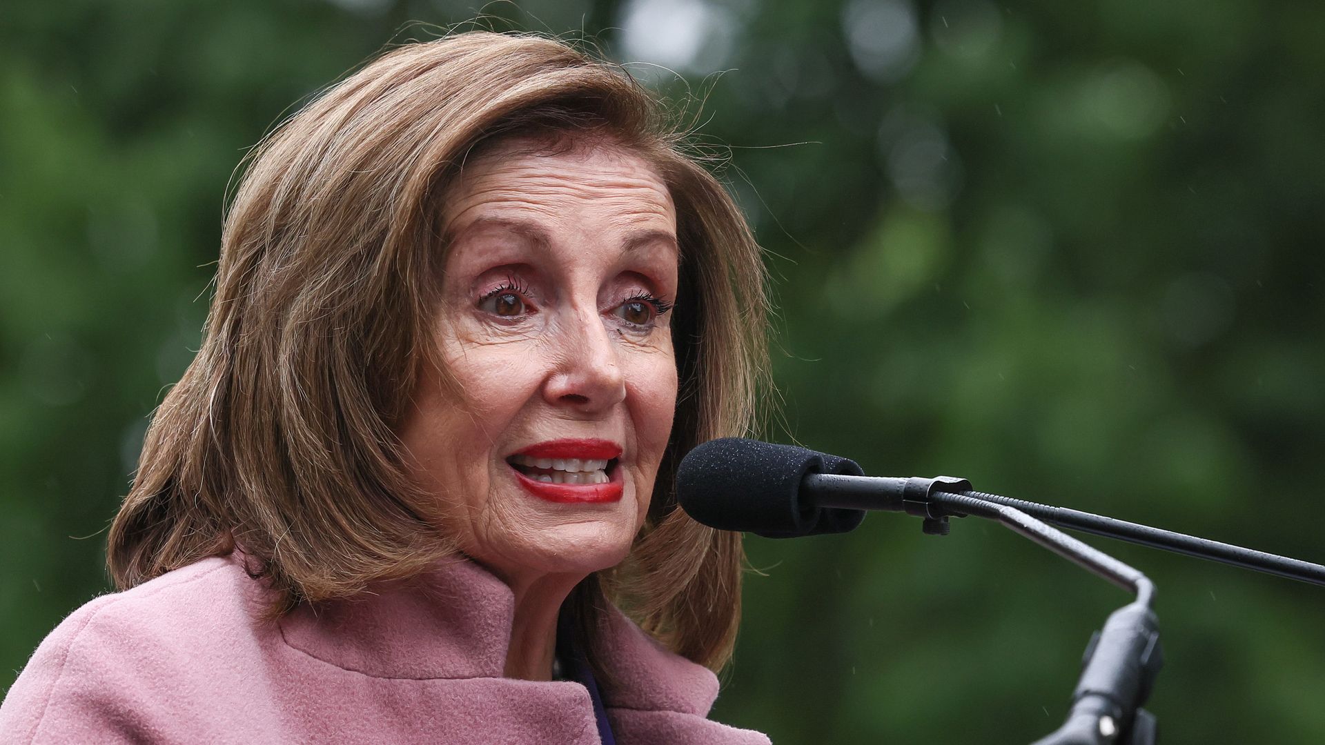 Nancy Pelosi wearing a pink coat and speaking into a microphone in front of trees.