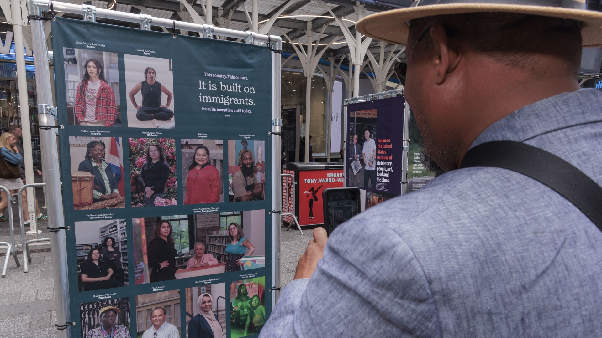 The back of man is shown as he takes a picture of a sign that features several immigrants for their contributions to the US