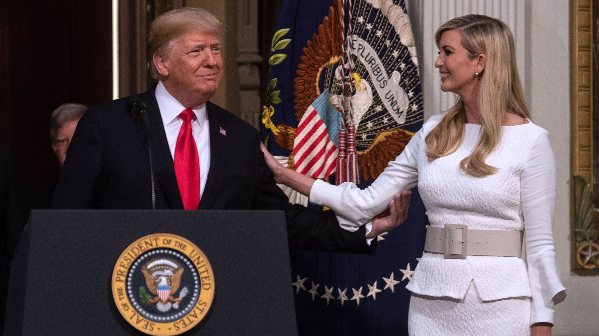 In this image, Ivanka stands next to Trump as he stands behind a podium. She smiles at him and places a hand on his shoulder.