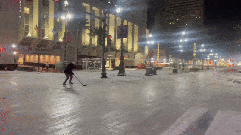 A person ice skates on Canal Street, pushing a puck around with a hockey stick.