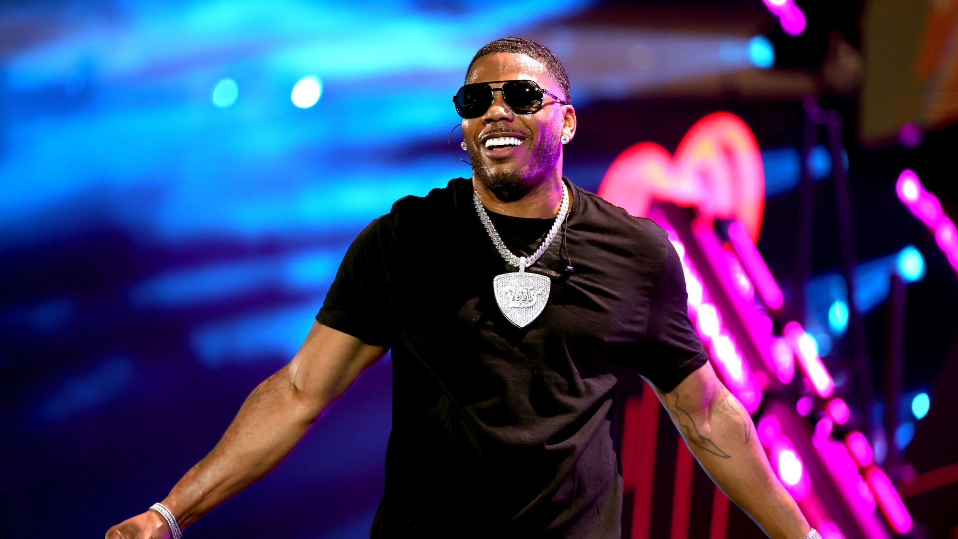 Rap star Nelly stands on stage with outstretched hands wearing sunglasses and a large necklace.